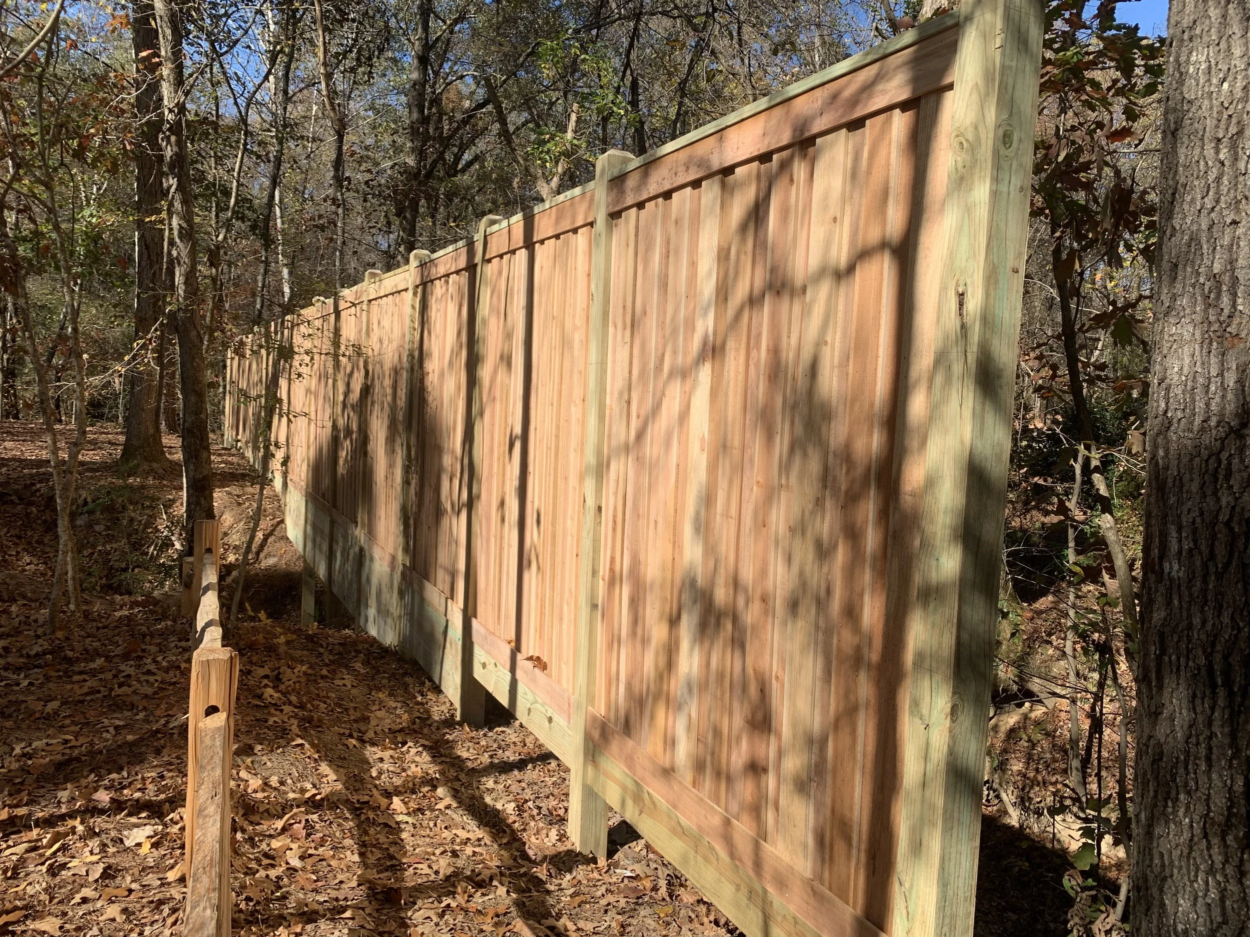 Wooden fence on a wooded hillside, shadows of trees cast on the fence, fallen leaves on the ground.