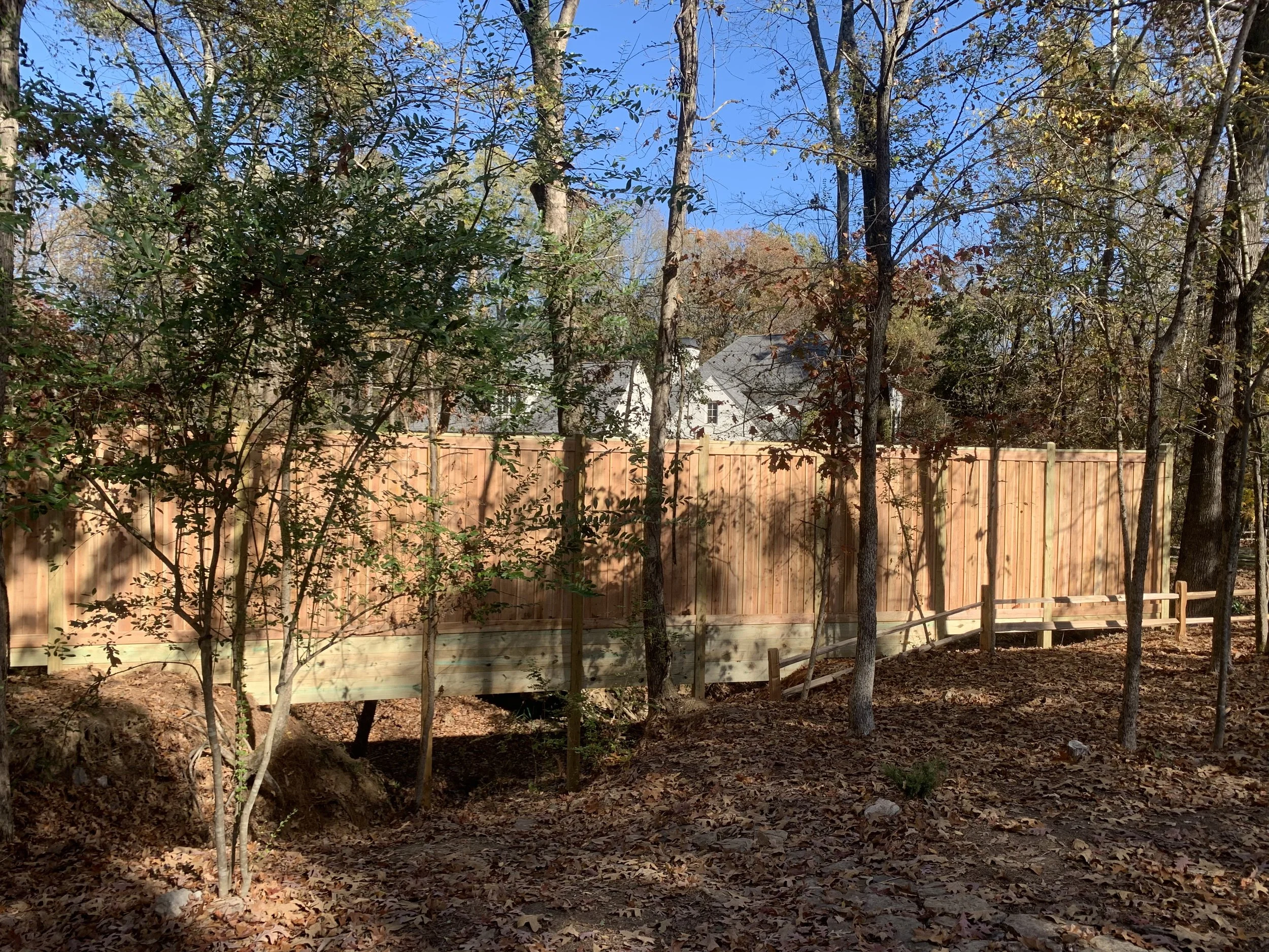 A wooden fence under construction in a backyard surrounded by trees with autumn leaves, with a house visible in the background on a clear, sunny day.
