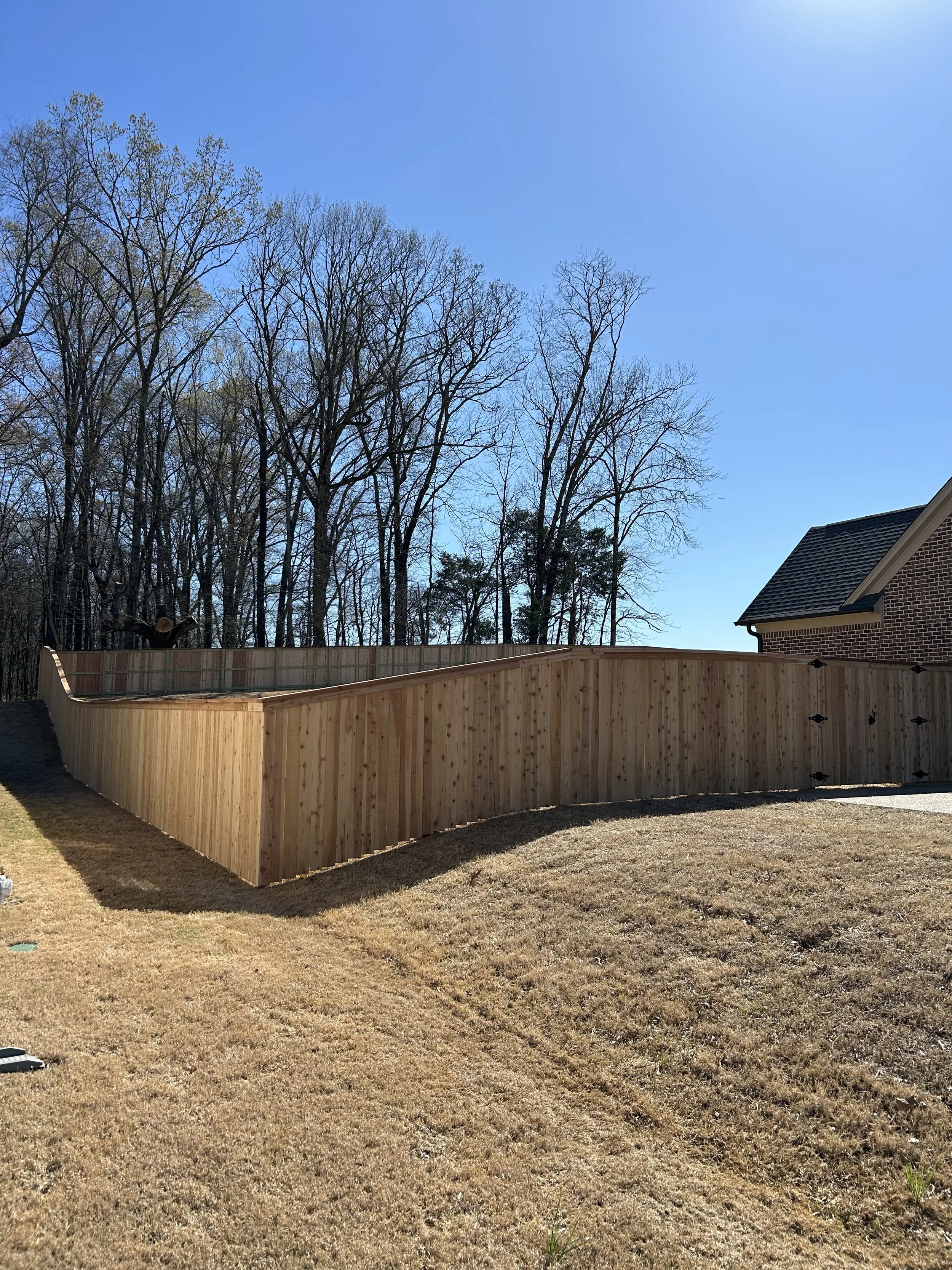 Newly built wooden privacy fence surrounding a backyard, with a house brick wall partially visible on the right, a grassy lawn in the front, and leafless trees in the background under a clear blue sky.