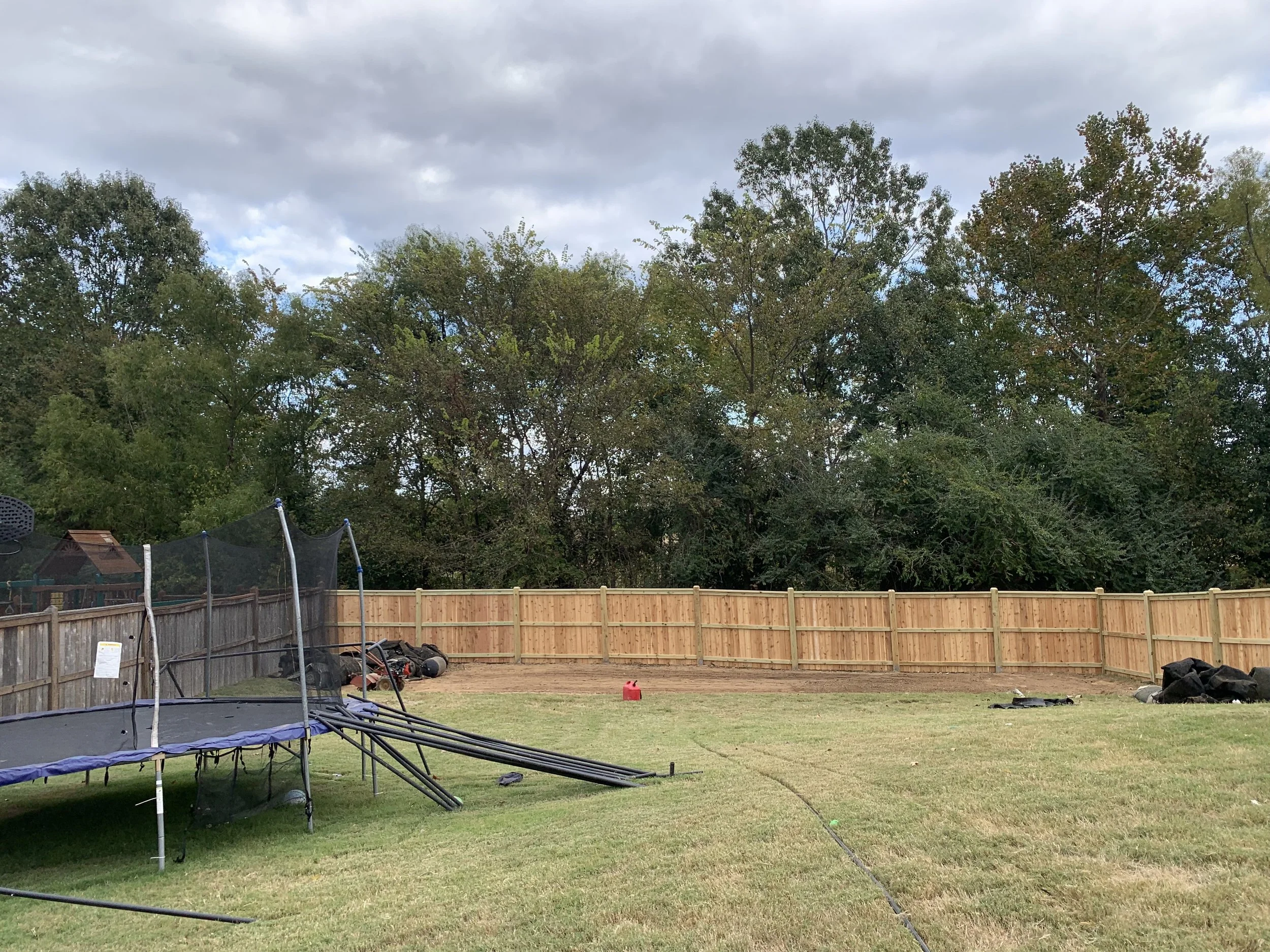 Backyard with a trampoline on the grass, surrounded by a wooden fence, with trees in the background and some yard tools and equipment on the ground.
