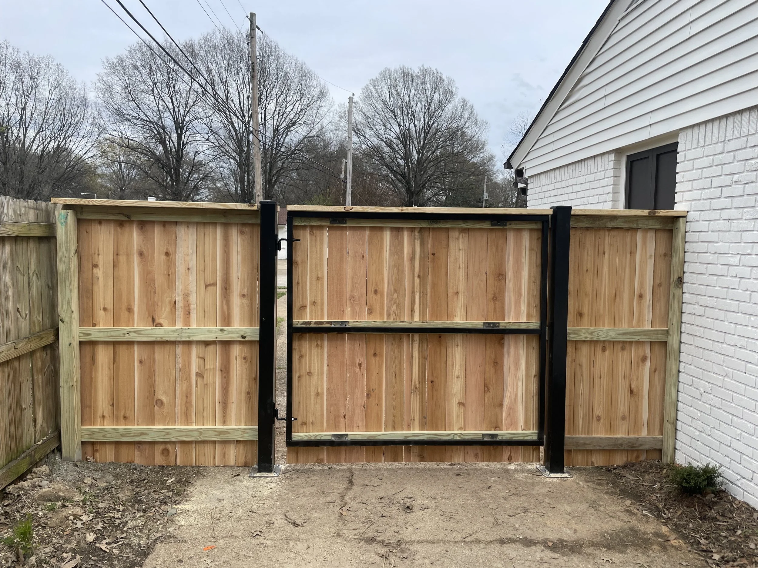 Newly installed wooden fence with black metal gate, white brick house on the right, and a background of leafless trees under a cloudy sky.