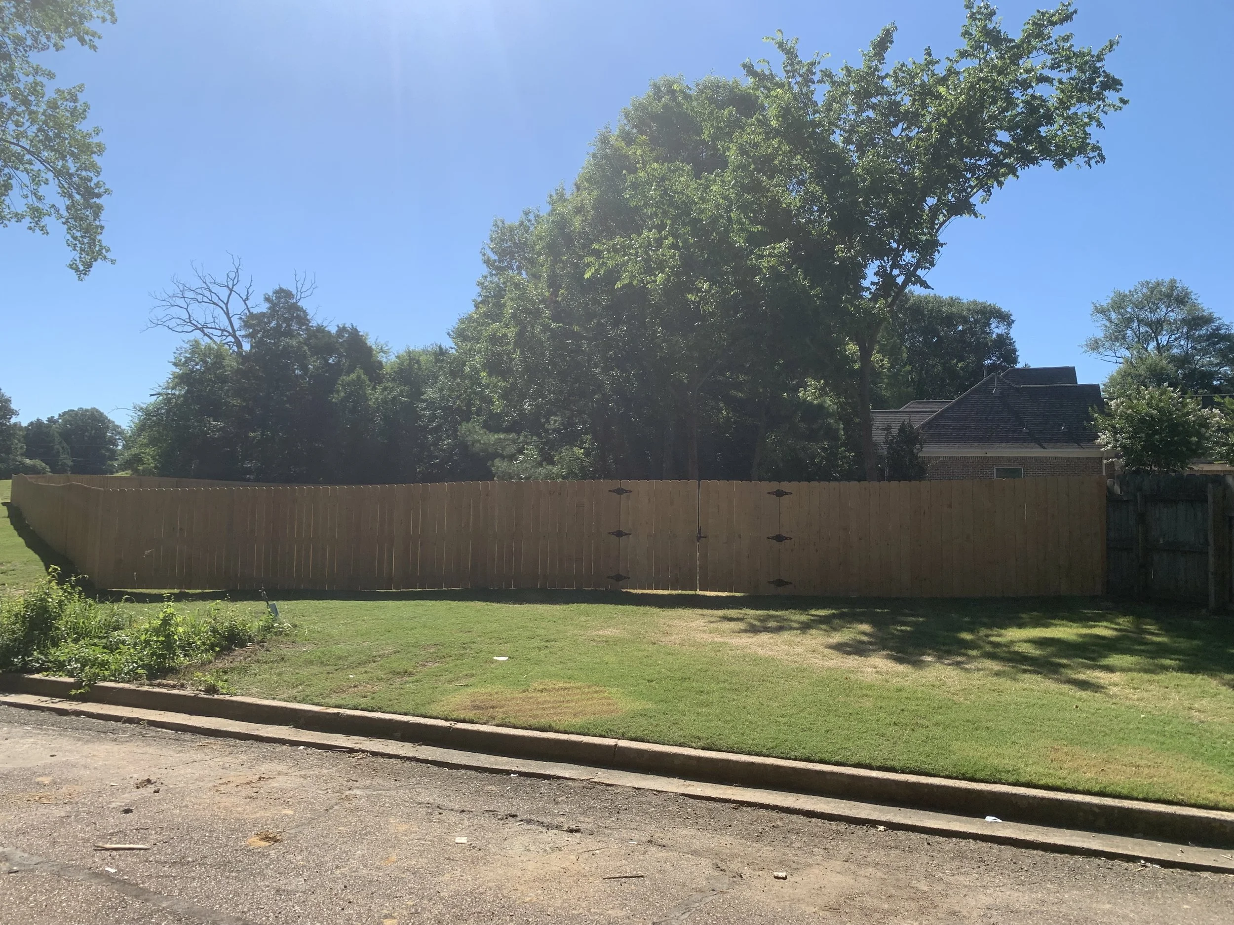 A freshly built wooden fence in a backyard, with a well-maintained grassy lawn, trees, and a house in the background, under a clear blue sky.