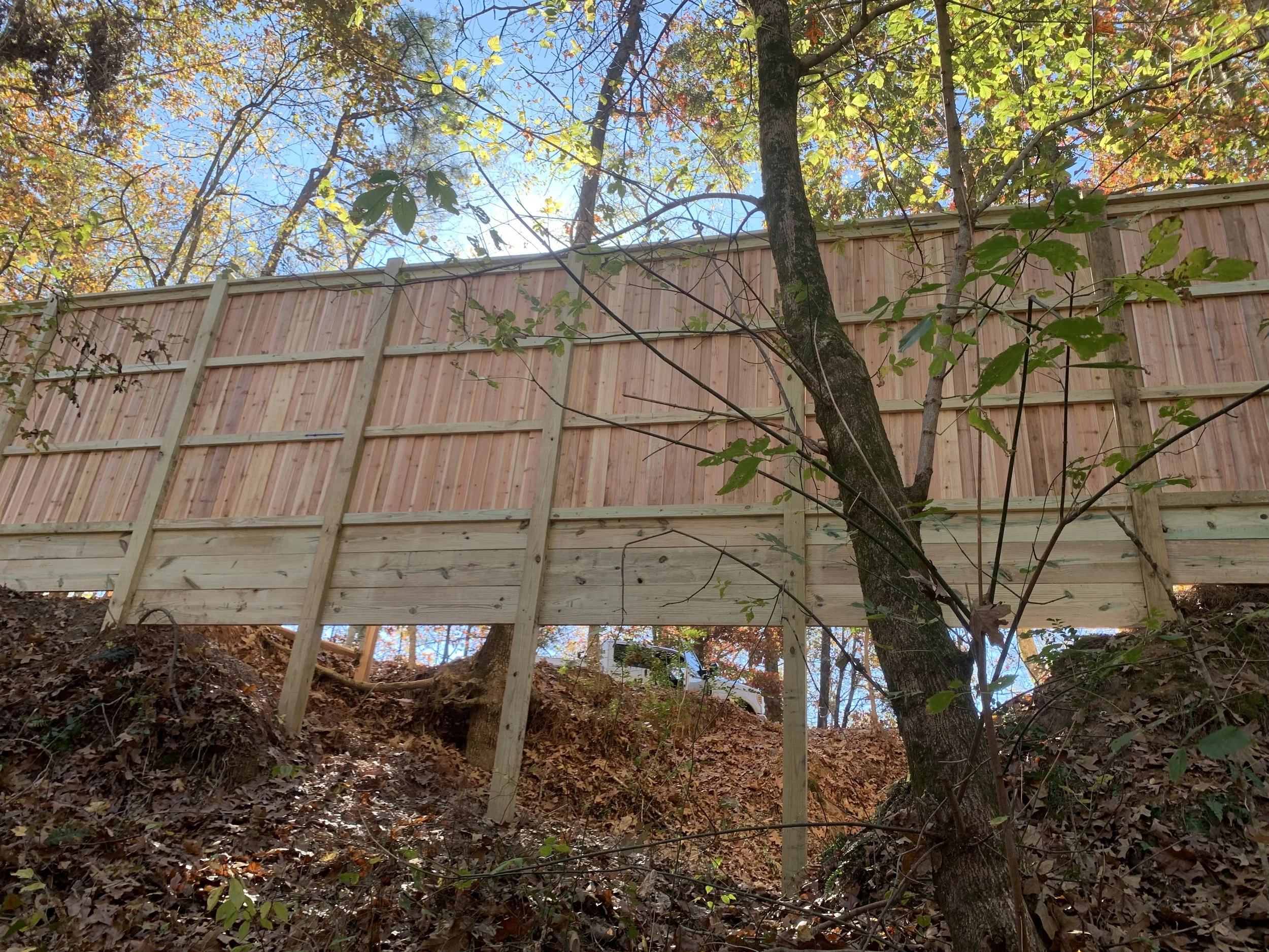 Wooden privacy fence being constructed on a hillside surrounded by trees with autumn leaves.