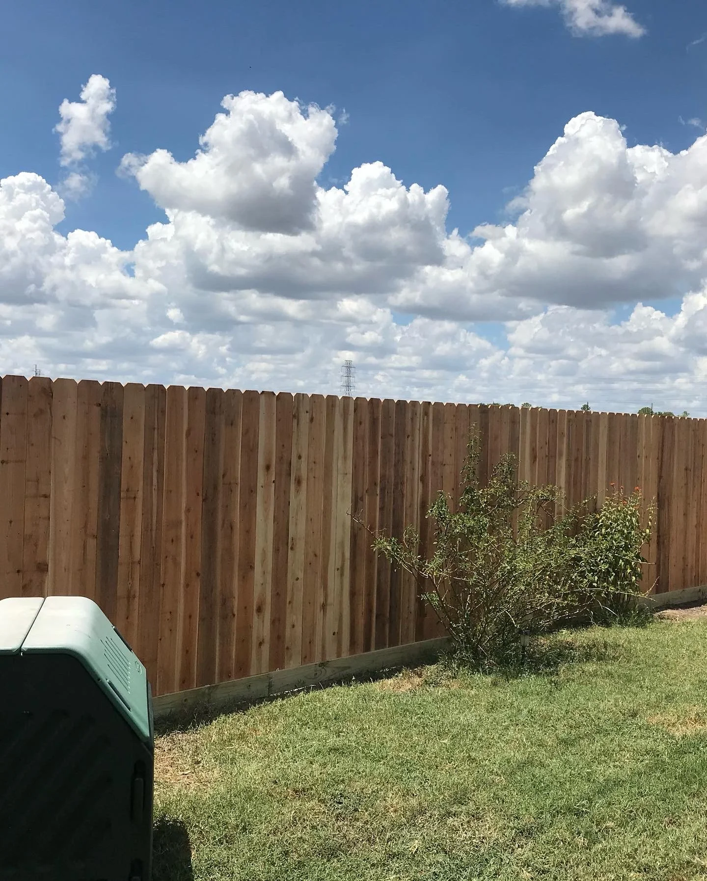 A backyard with a brown wooden fence, green grass, a small bush, cloudy sky with white and gray clouds, and a part of a light-colored outdoor appliance in the lower left corner.
