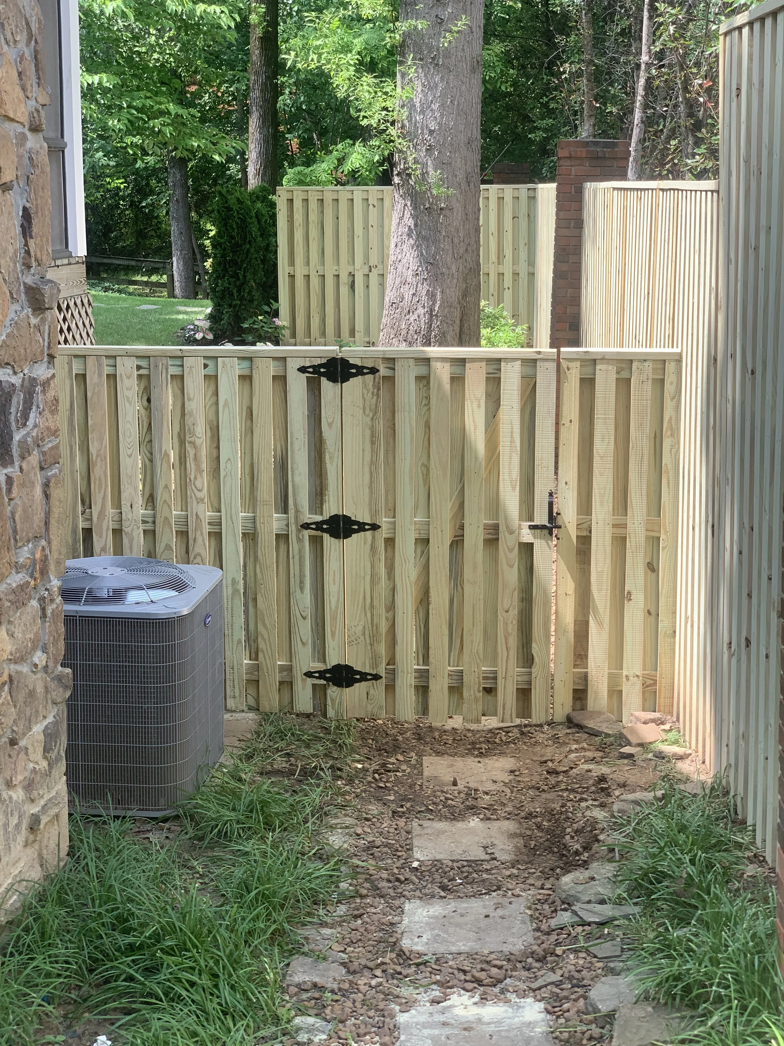A small backyard area enclosed by a new wooden fence with a gate, an air conditioning unit on the left, and a dirt pathway with stepping stones leading to the gate. There are trees and greenery in the background.