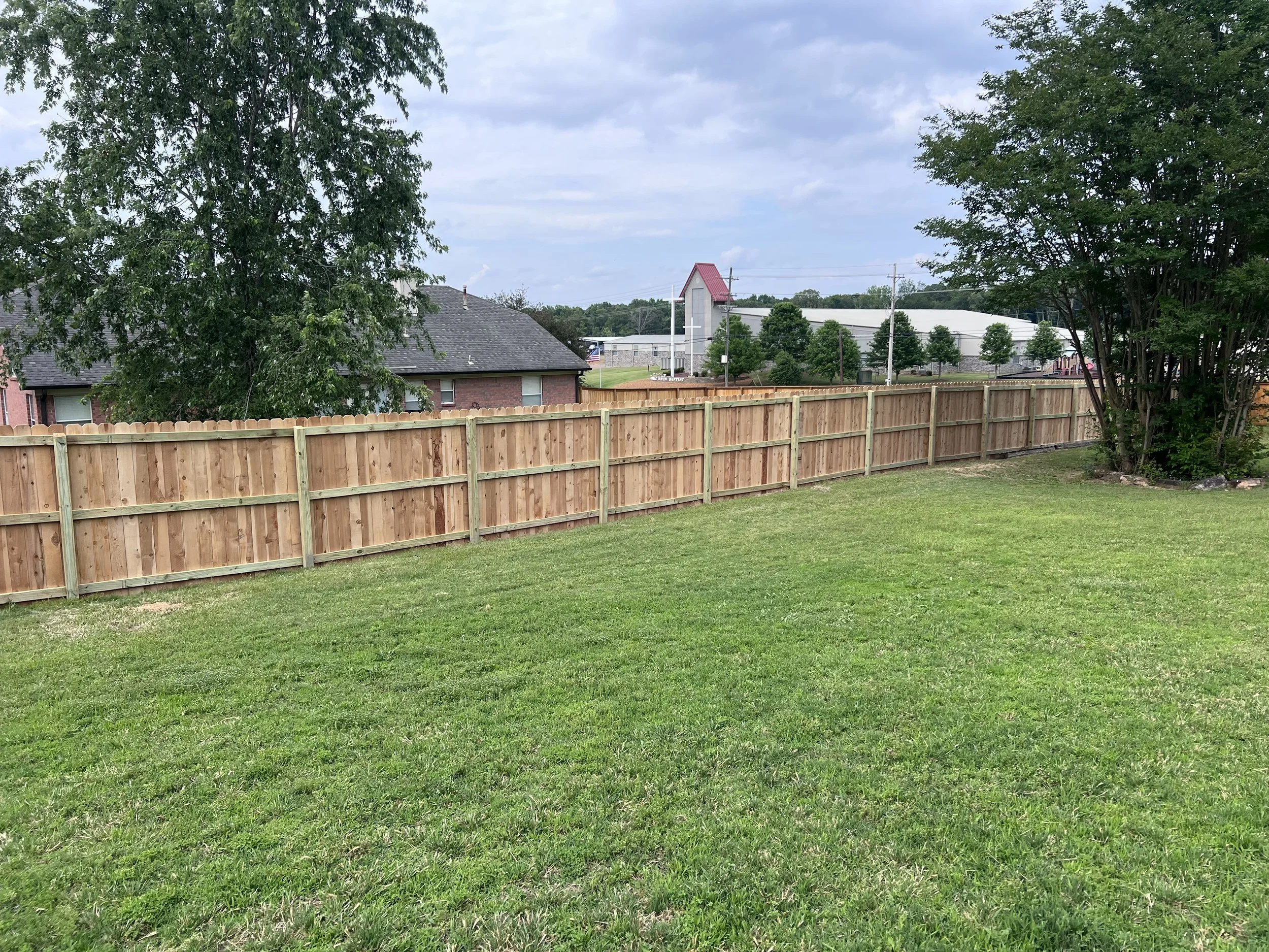 A backyard with a green lawn, a wooden fence, trees, and a building with a church tower in the background.