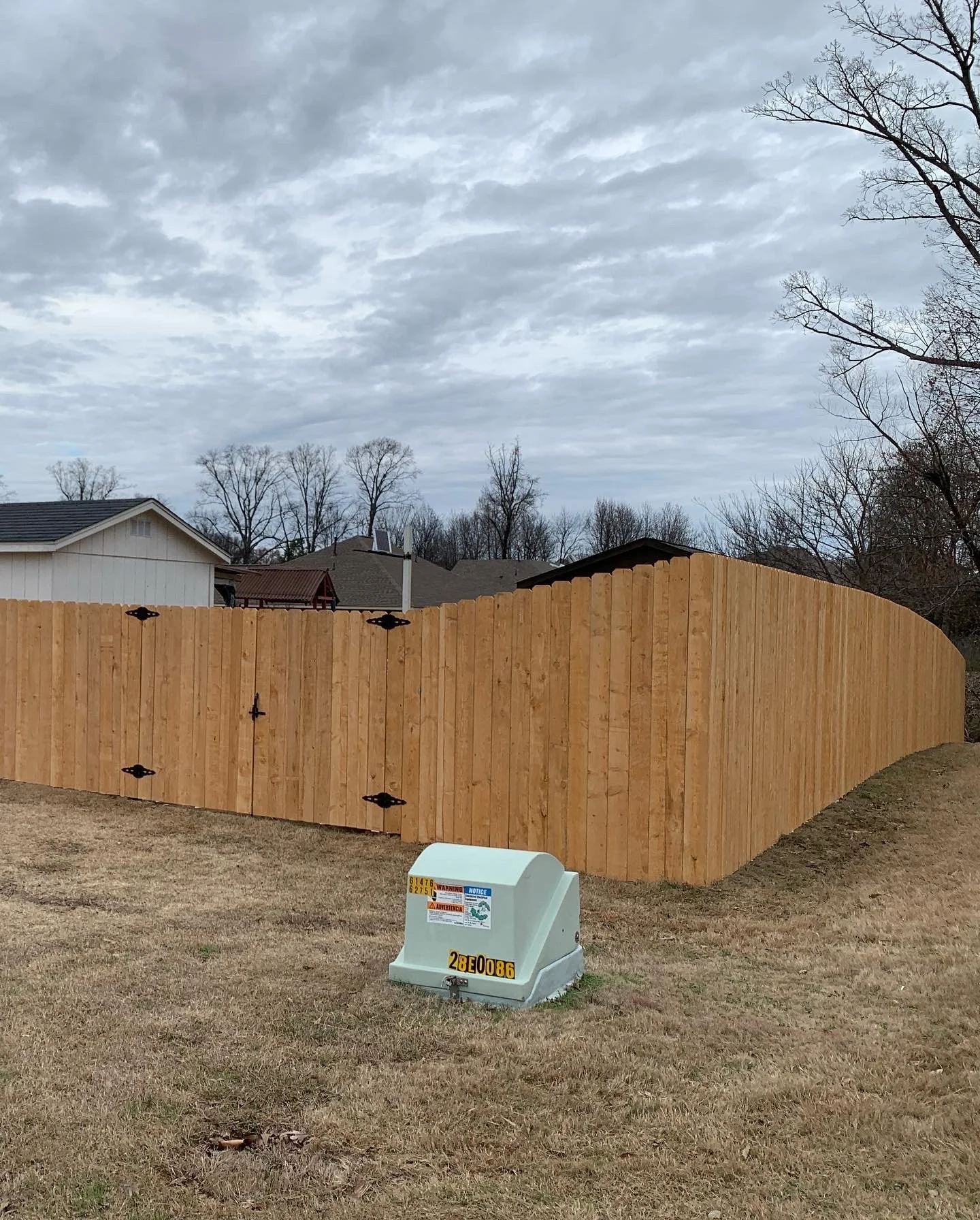 A newly installed wooden privacy fence in a suburban backyard with a cloudy sky overhead. There is a utility box on the grass in the foreground.
