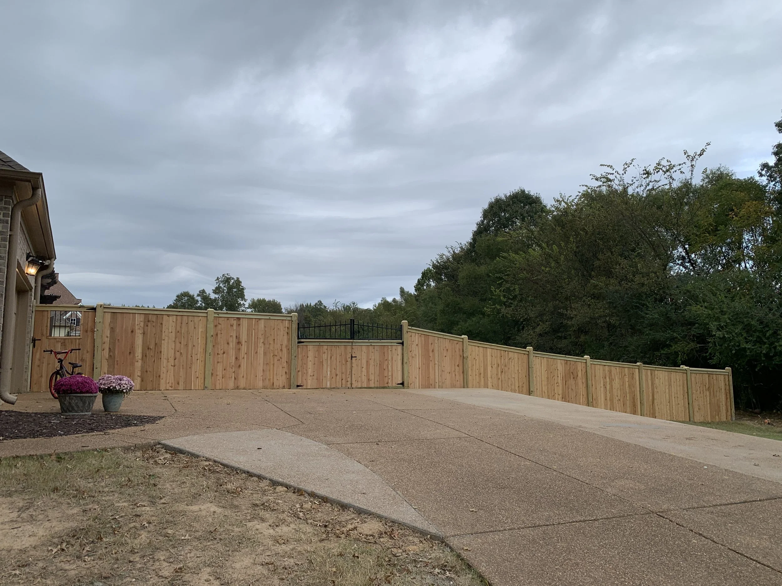 Residential backyard with a newly installed wooden fence, cloudy sky, and a sloped driveway. Two flower pots with pink flowers and a child's bicycle are near the house on the left side of the image.
