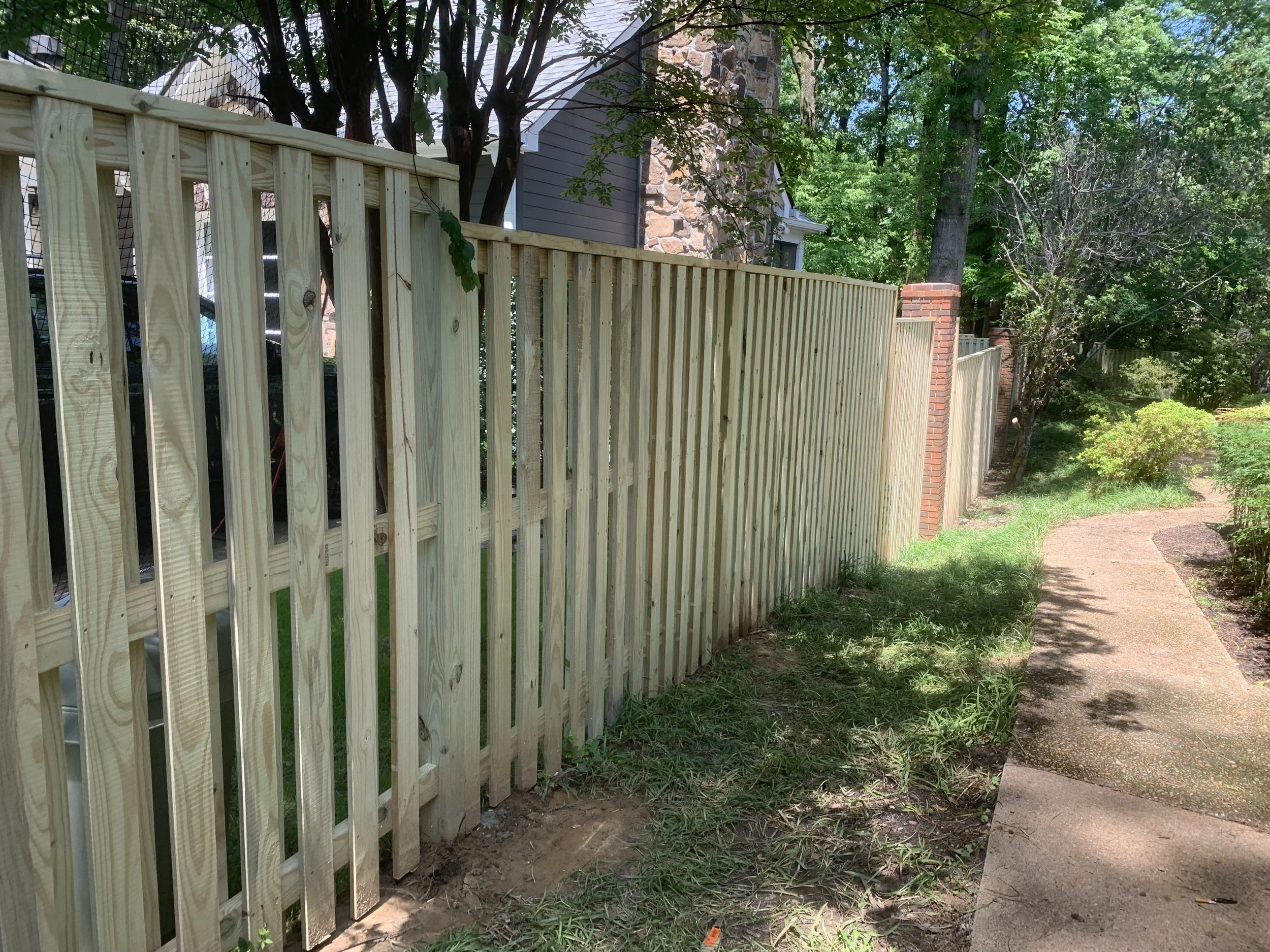 A newly constructed wooden fence running alongside a sidewalk in a residential area, with trees and shrubs in the background.