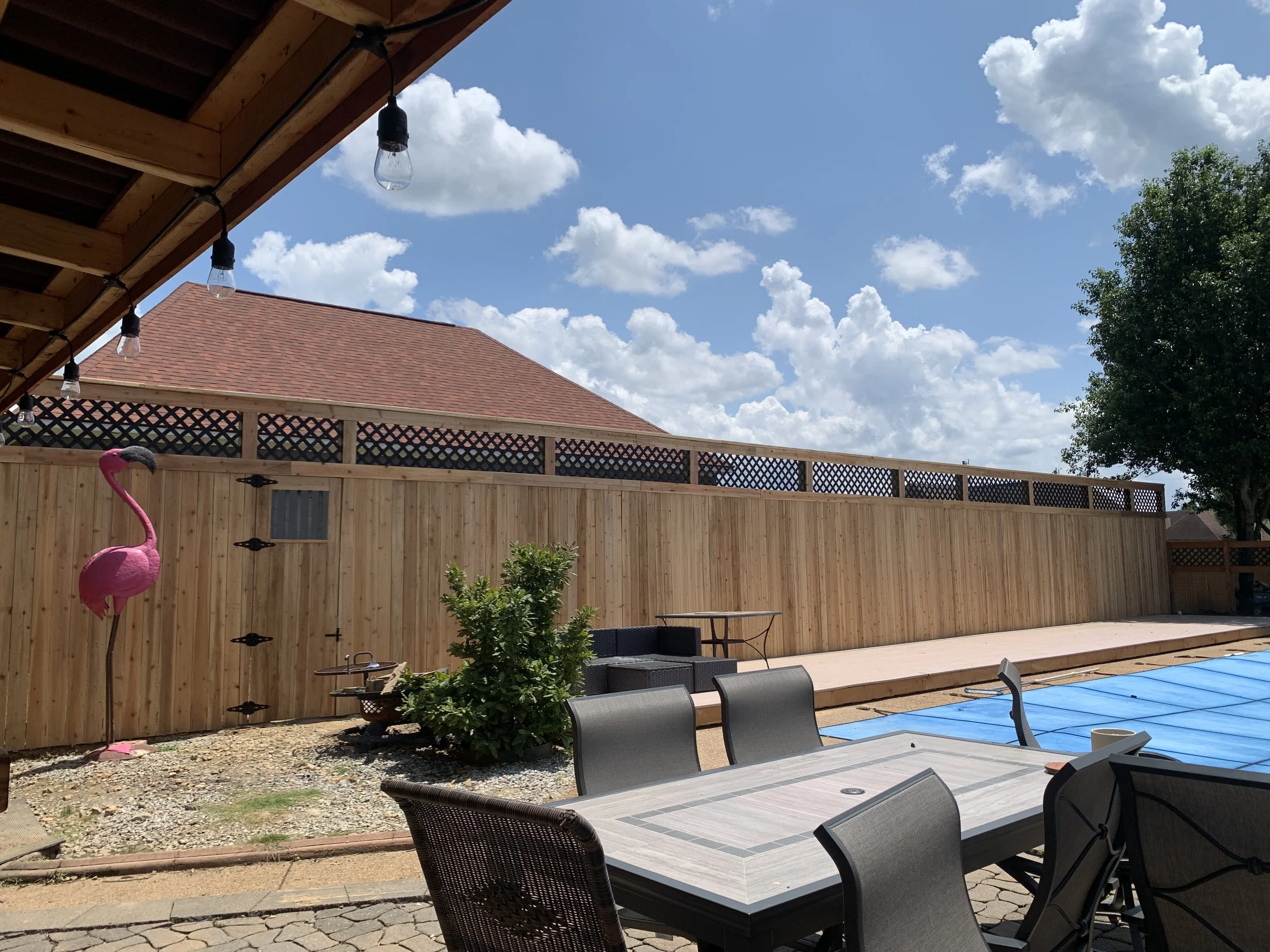 Backyard patio with outdoor furniture, a wooden privacy fence, a pink flamingo garden ornament, and a pool covered with a blue tarp. The sky is partly cloudy.