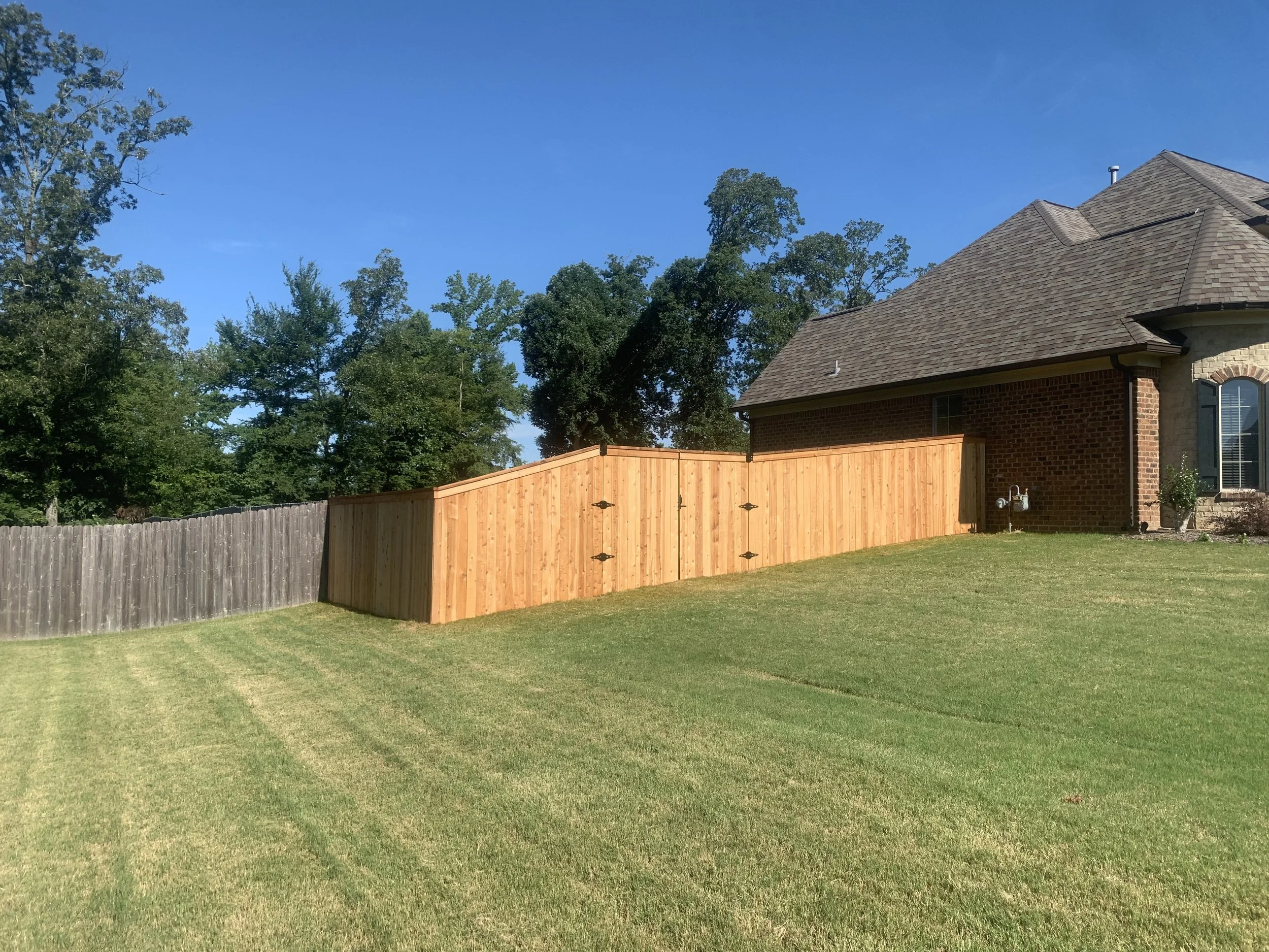 New wooden privacy fence installed next to house with brick exterior, surrounded by a well-maintained green lawn and trees under a clear blue sky.