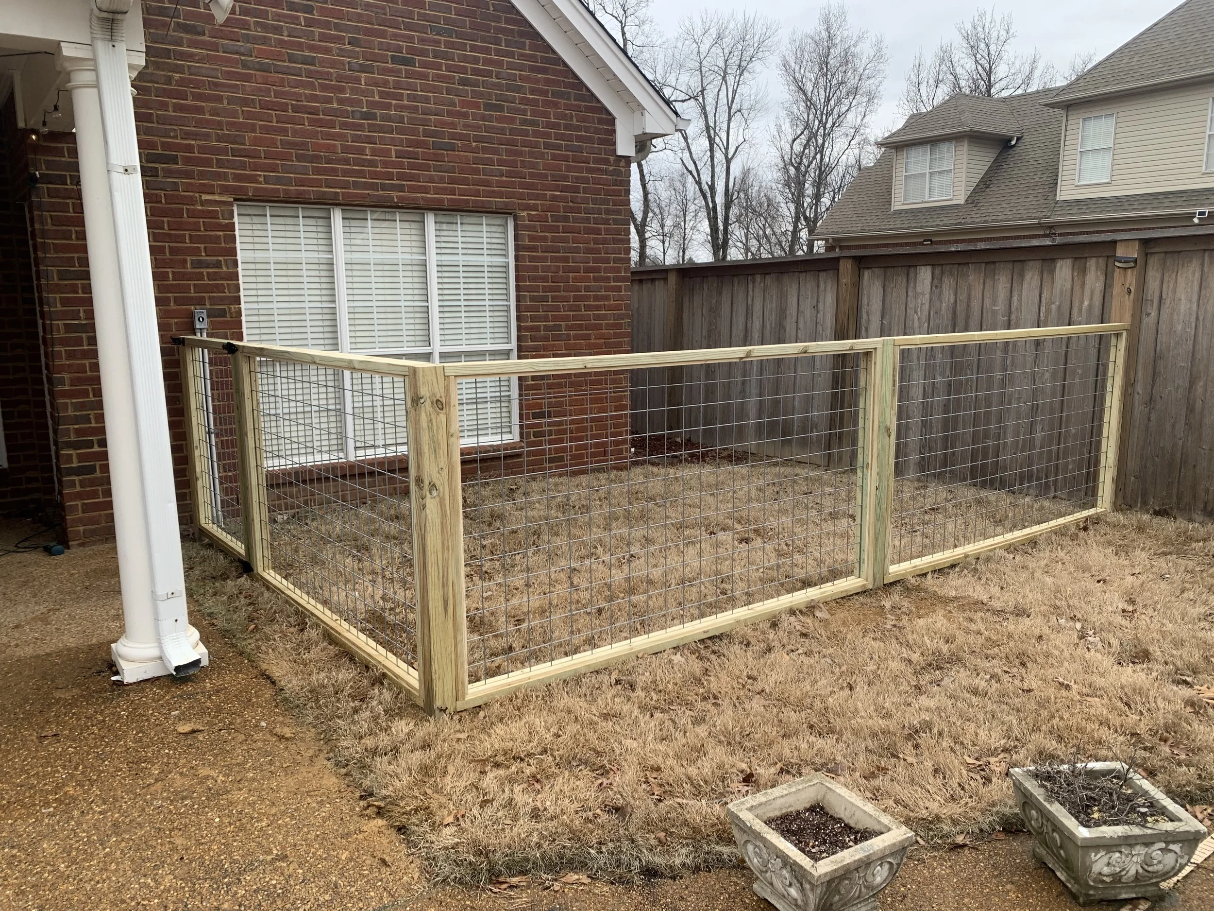 A newly constructed wooden and wire fence enclosure in a backyard, adjacent to a brick house with a window, and a concrete patio area with two empty planters.