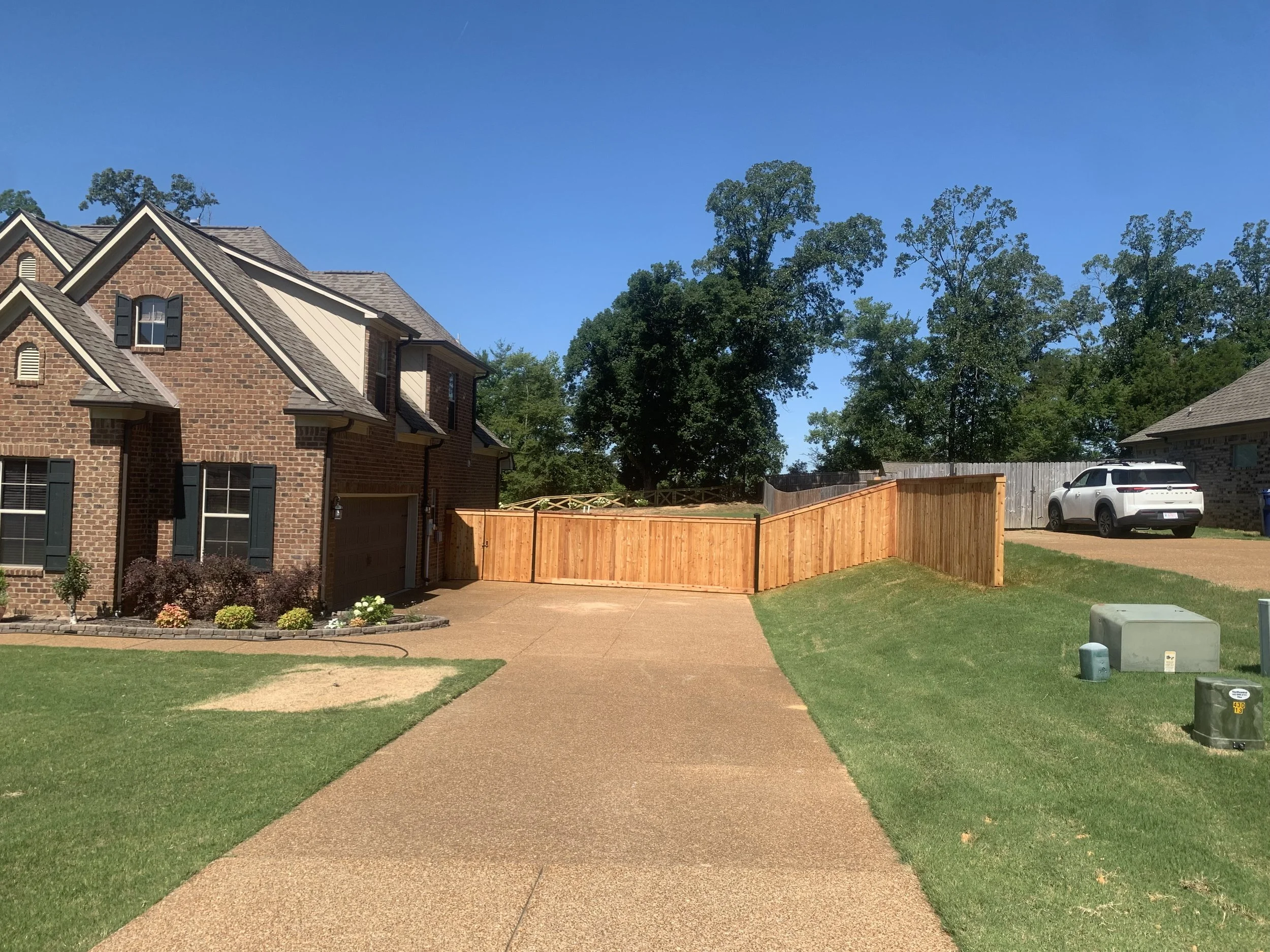 A suburban house with a brick exterior and a brown garage, surrounded by a well-maintained yard. A new wooden fence extends from the house across the yard, and a white SUV is parked on the driveway in the background. Bright blue sky and green trees a