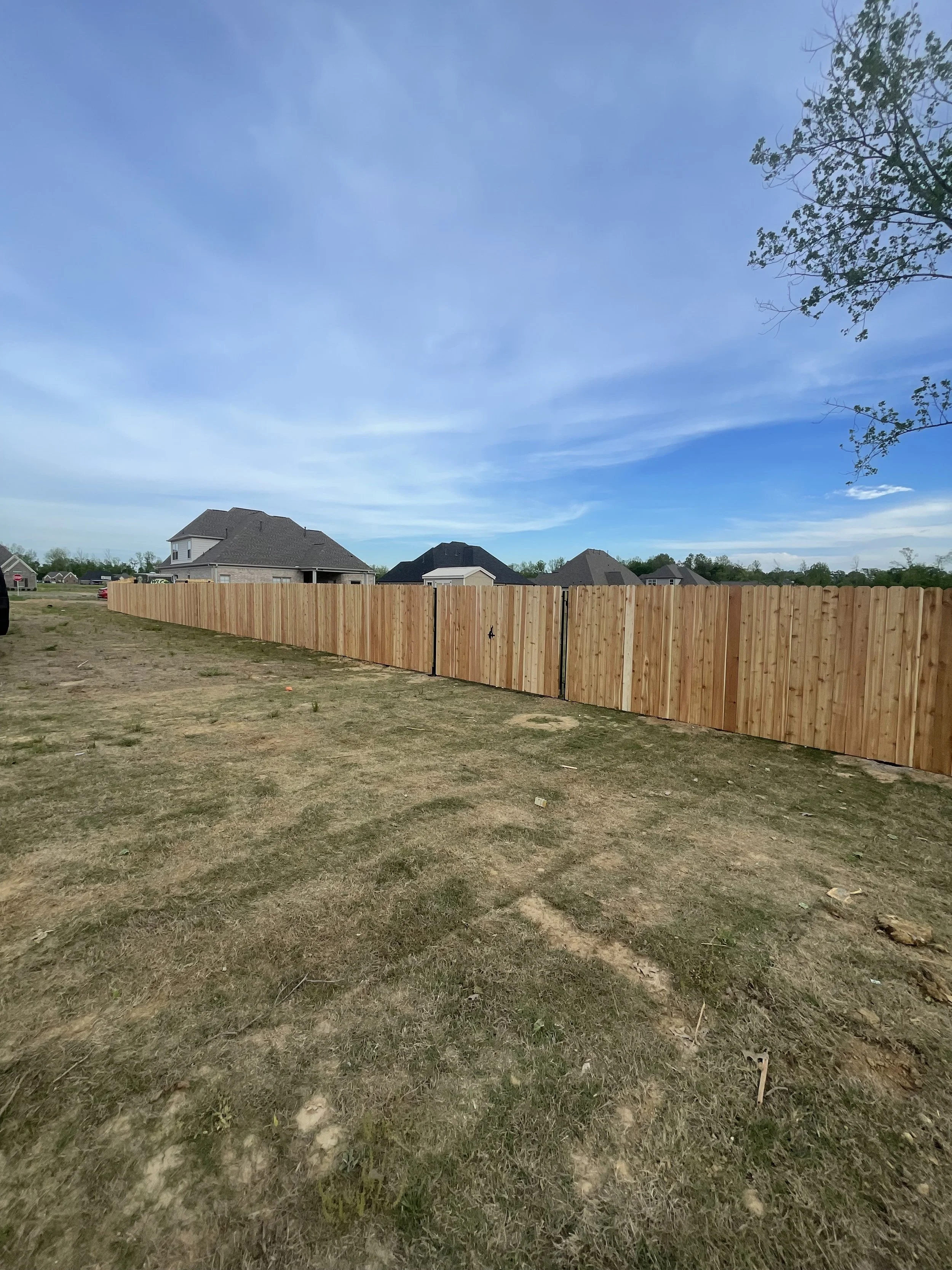 A newly built wooden fence in a residential yard with several houses in the background and a partly cloudy sky.