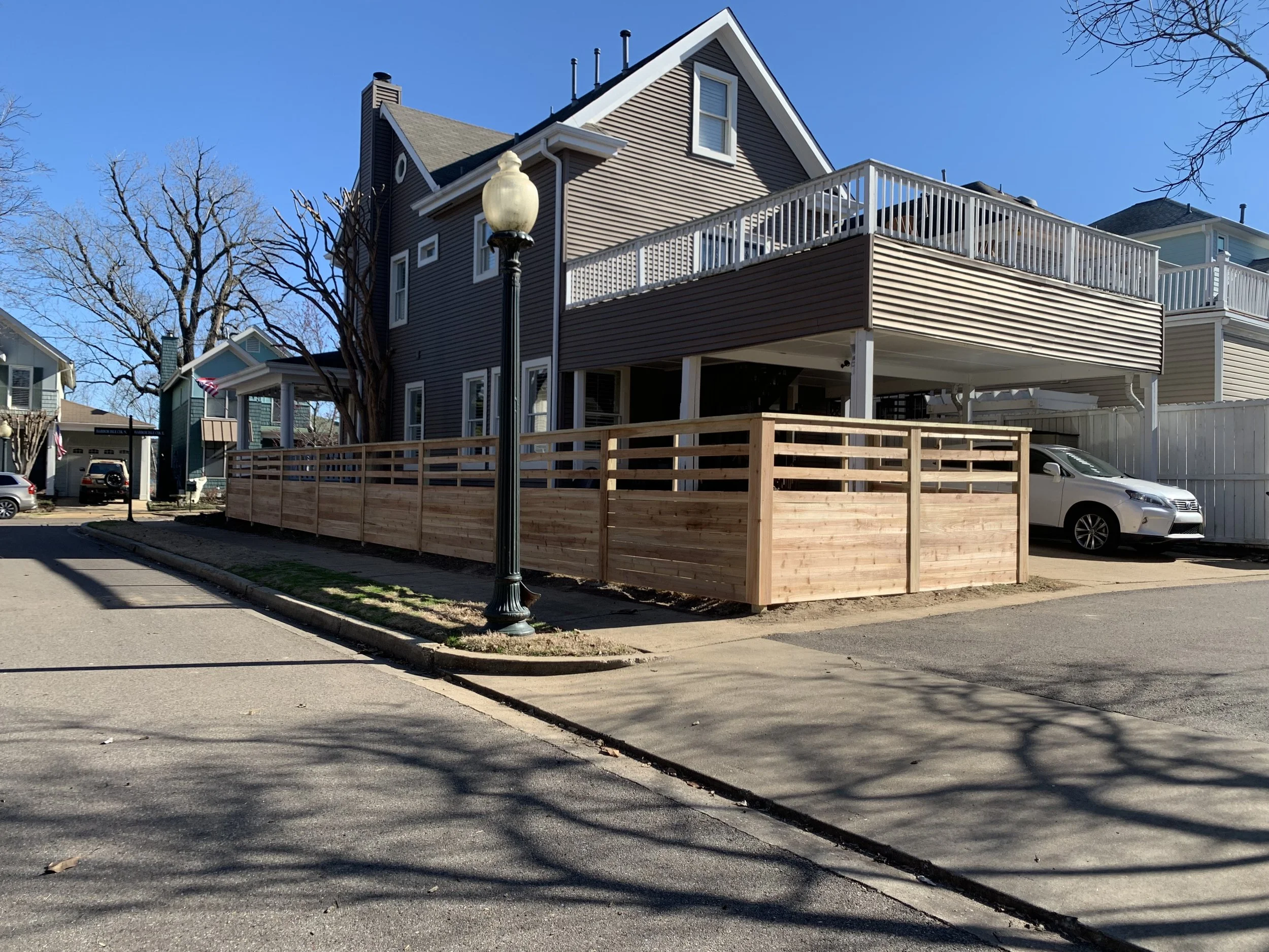 Residential house with a wooden fence and an elevated deck, parked white car, street lamp, leafless trees, and neighboring houses under clear blue sky.