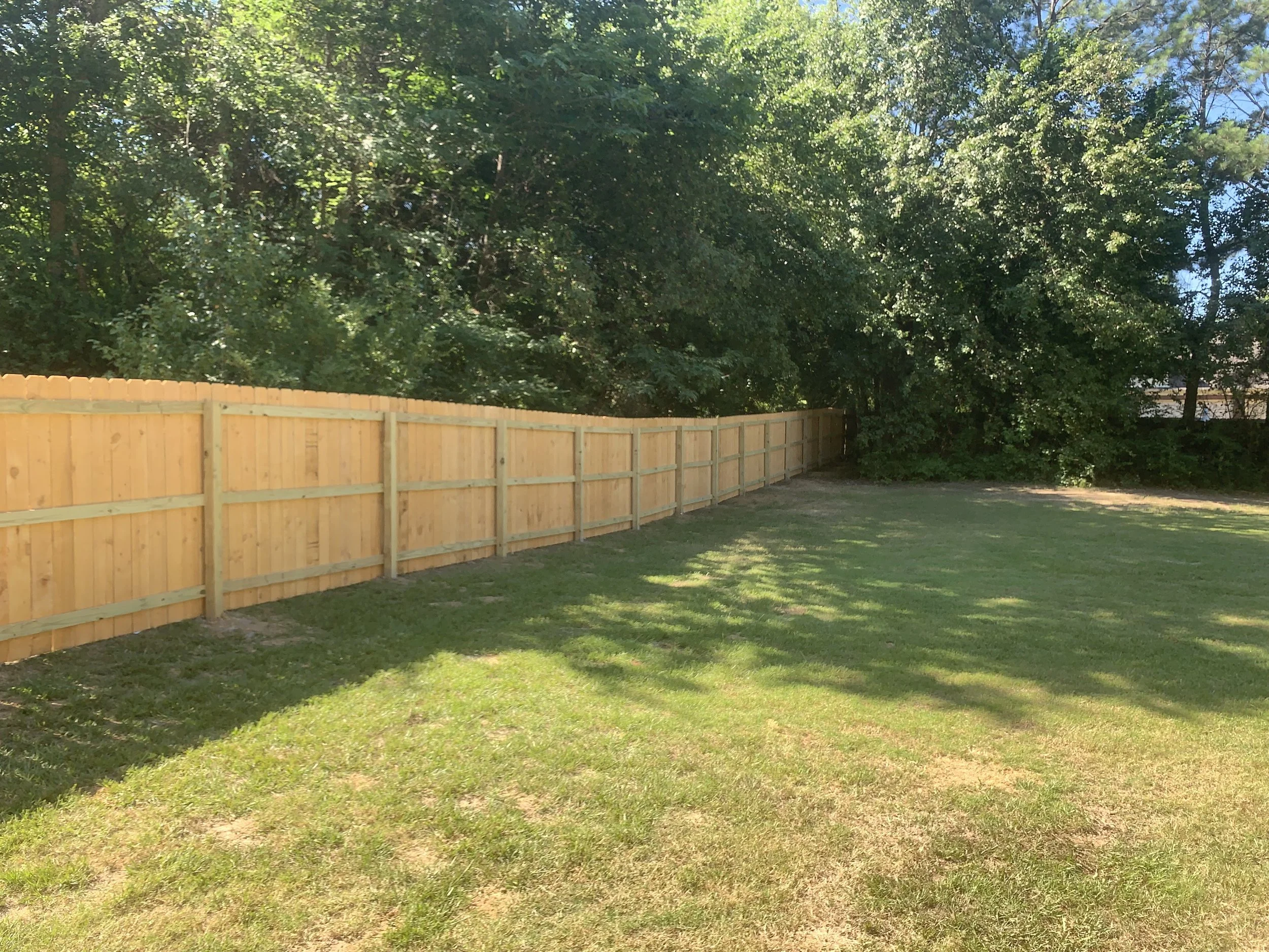 Freshly installed wooden privacy fence running along a grassy backyard with trees in the background.