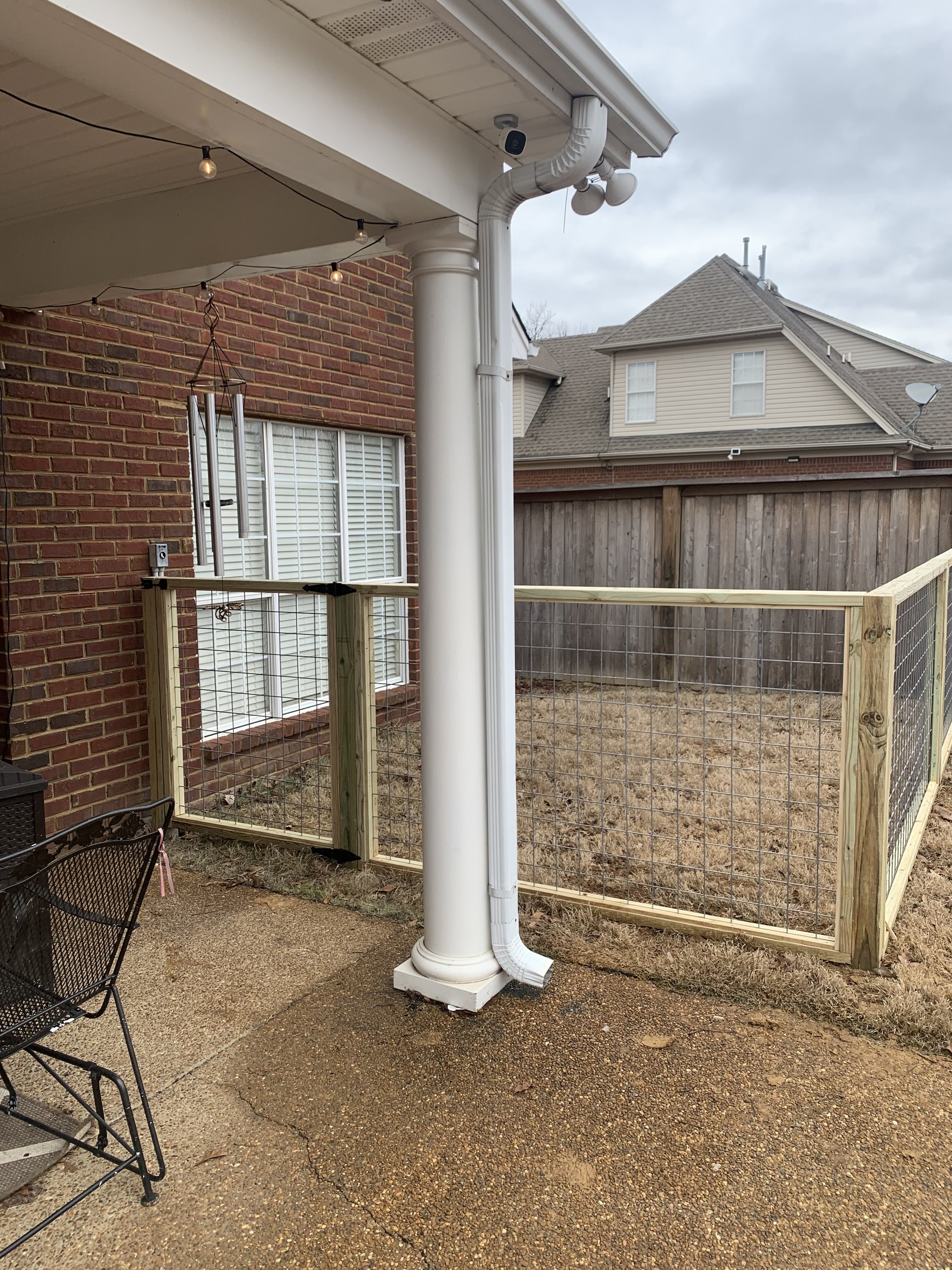 Backyard patio with a small fenced area, brick house with white window blinds, and a chimney with wind chimes hanging. Overcast sky.
