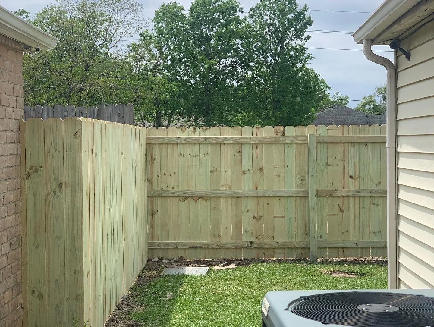 Backyard enclosed by new wooden fence, with a small grassy area and a part of an air conditioning unit visible in the foreground.
