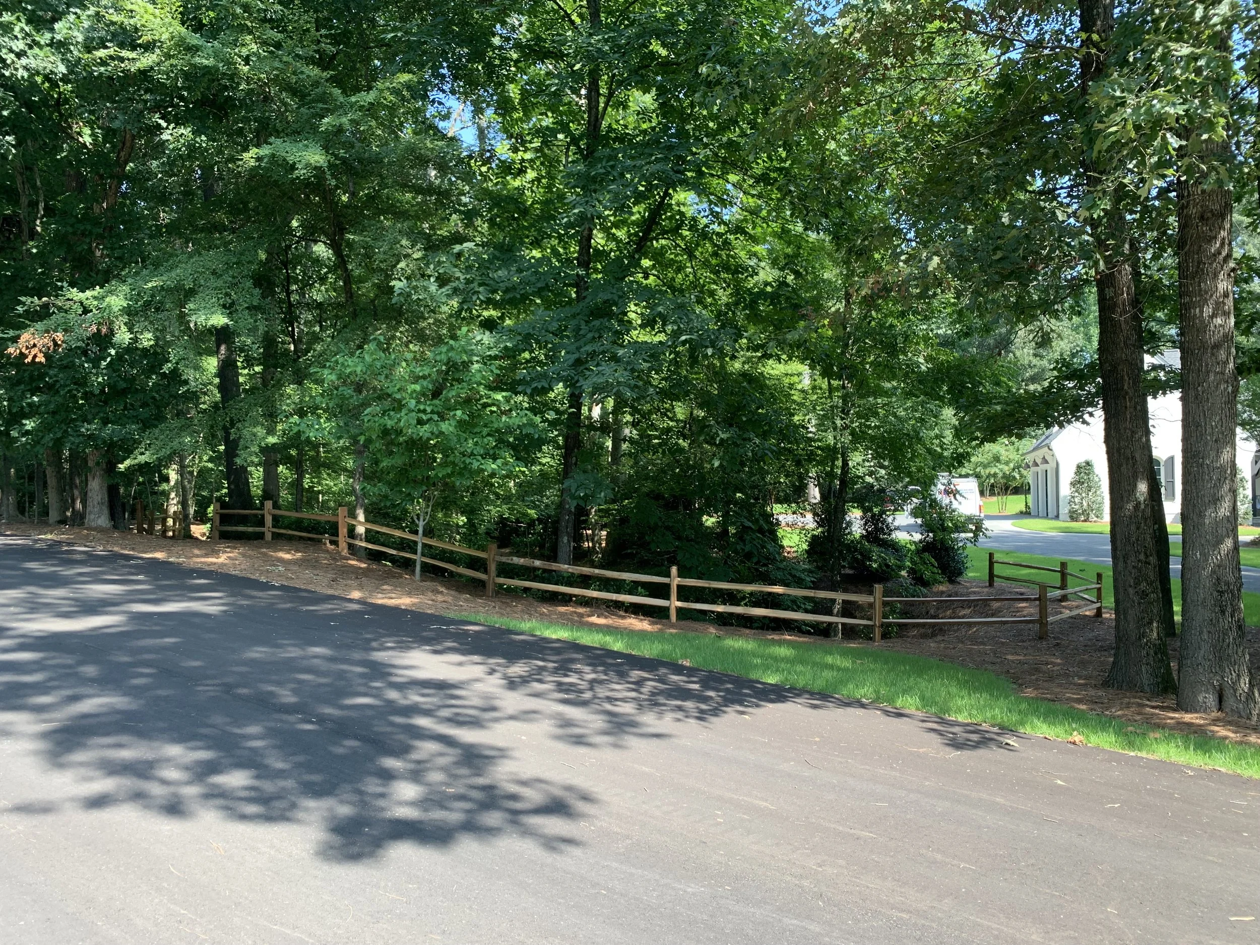 A paved road runs alongside a wooded area with a wooden fence, green grass, and trees. In the background, there are residential houses and a driveway.