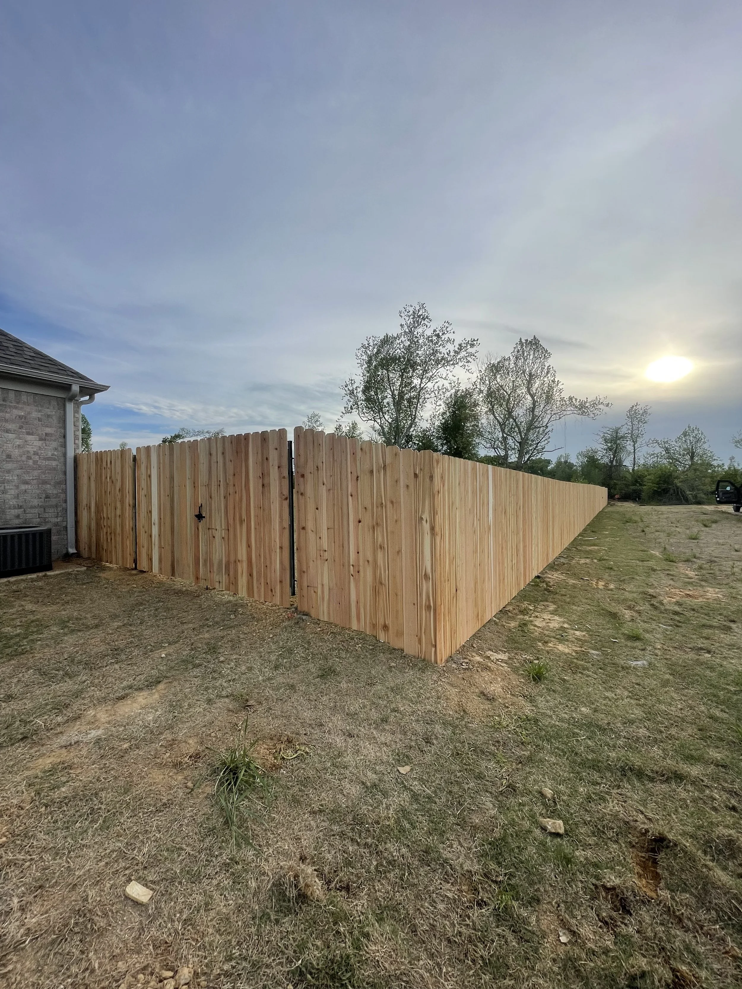 Newly constructed wooden fence surrounding backyard with a house visible on the left, trees in the background, and a setting sun.