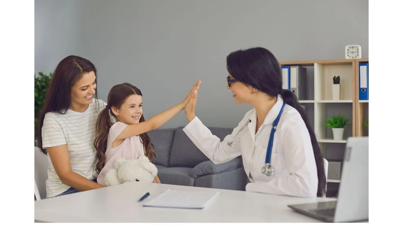 A young girl giving a high five to a female doctor in an office with her mother, with a teddy bear and a notebook on the desk.