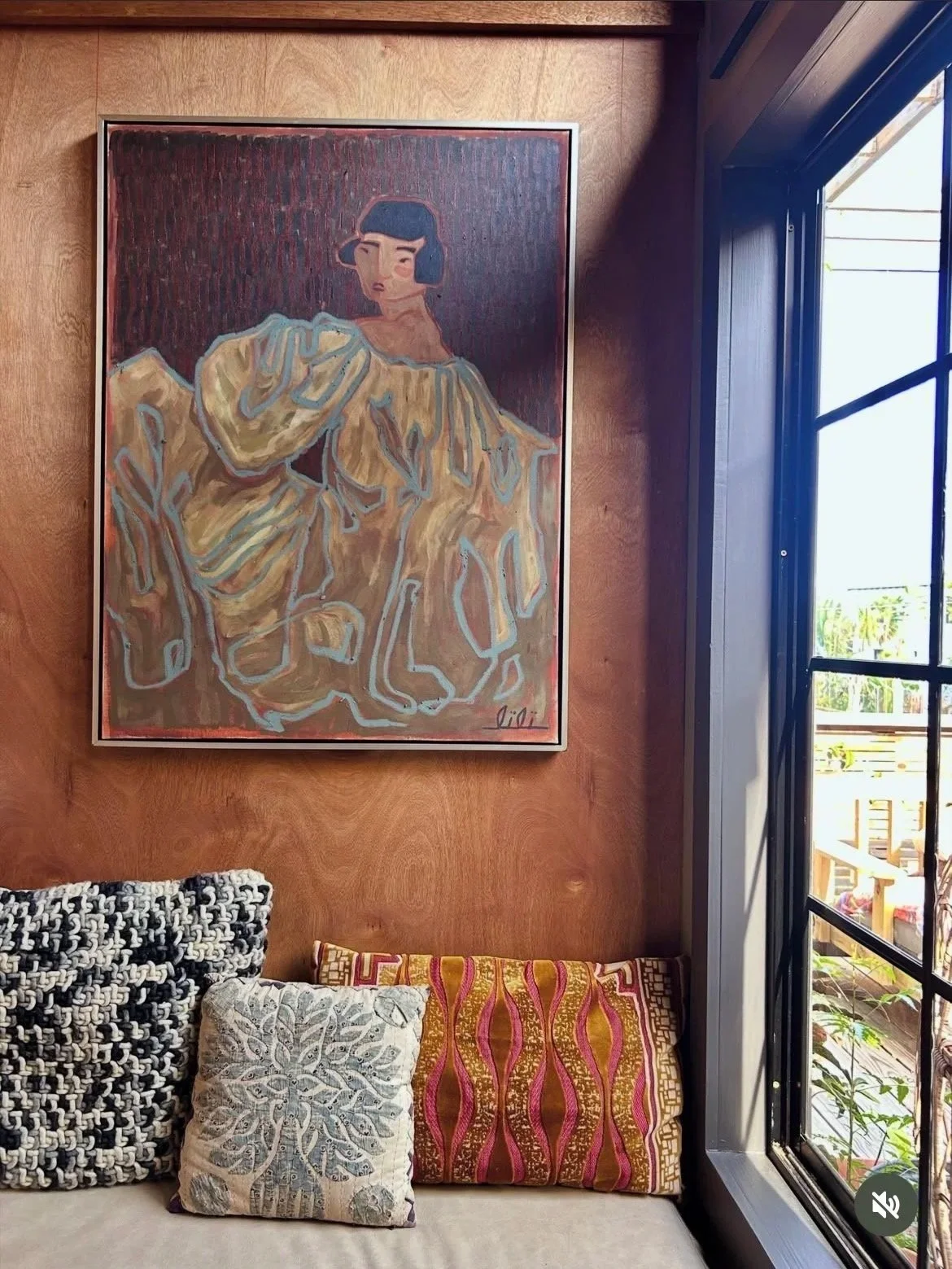 An original painting of a woman with short black hair, wearing a flowing yellow dress with blue outlines by artist Lili Dauphinee. The painting is framed and hangs on a wood-clad wall with well designed interior decorating.