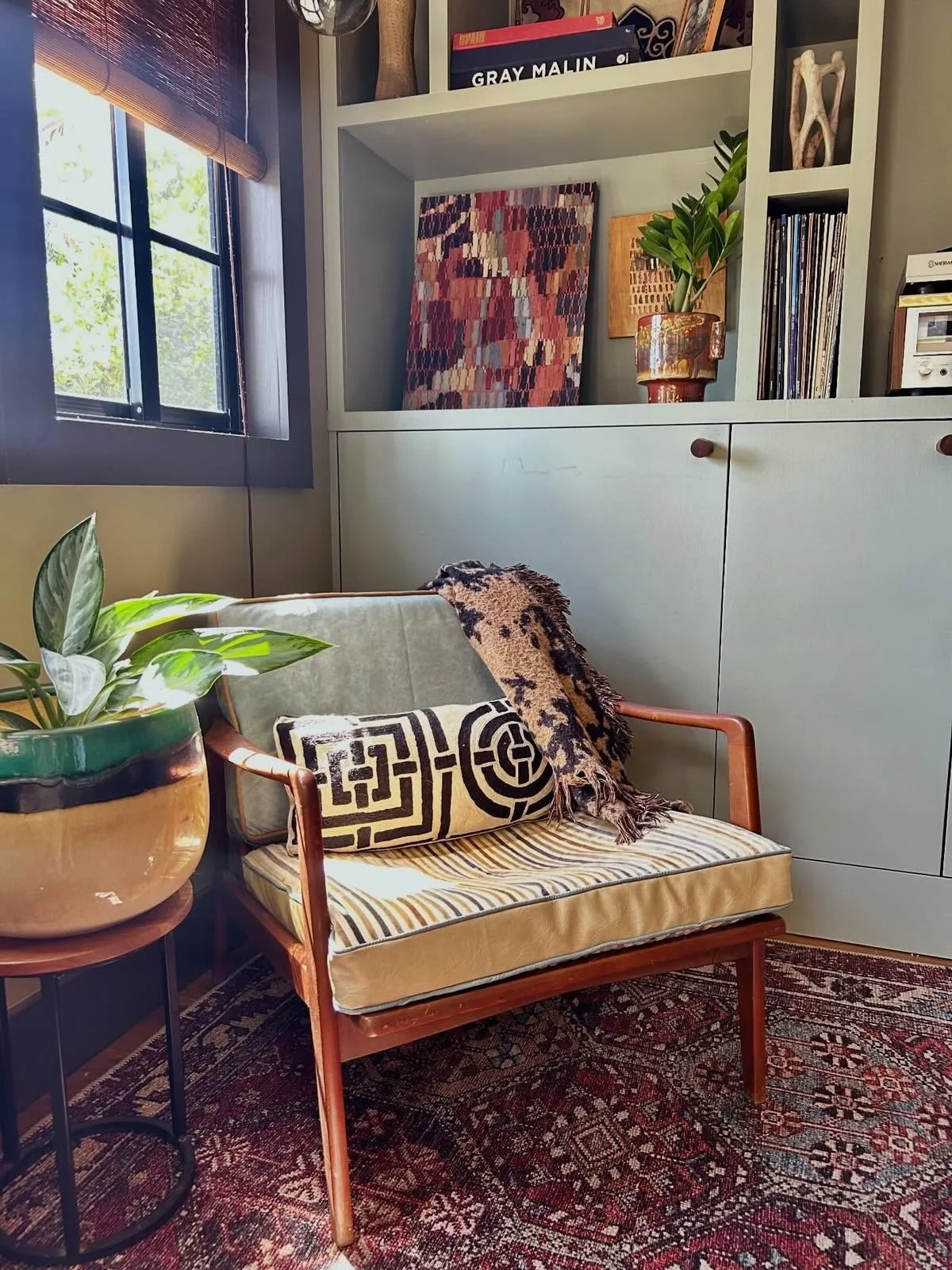 A cozy corner with a wooden armchair featuring a patterned pillow and a blanket, next to a large potted plant on a small round table, near a window with blinds. A white bookshelf with an original painting by artist Lili Dauphinee