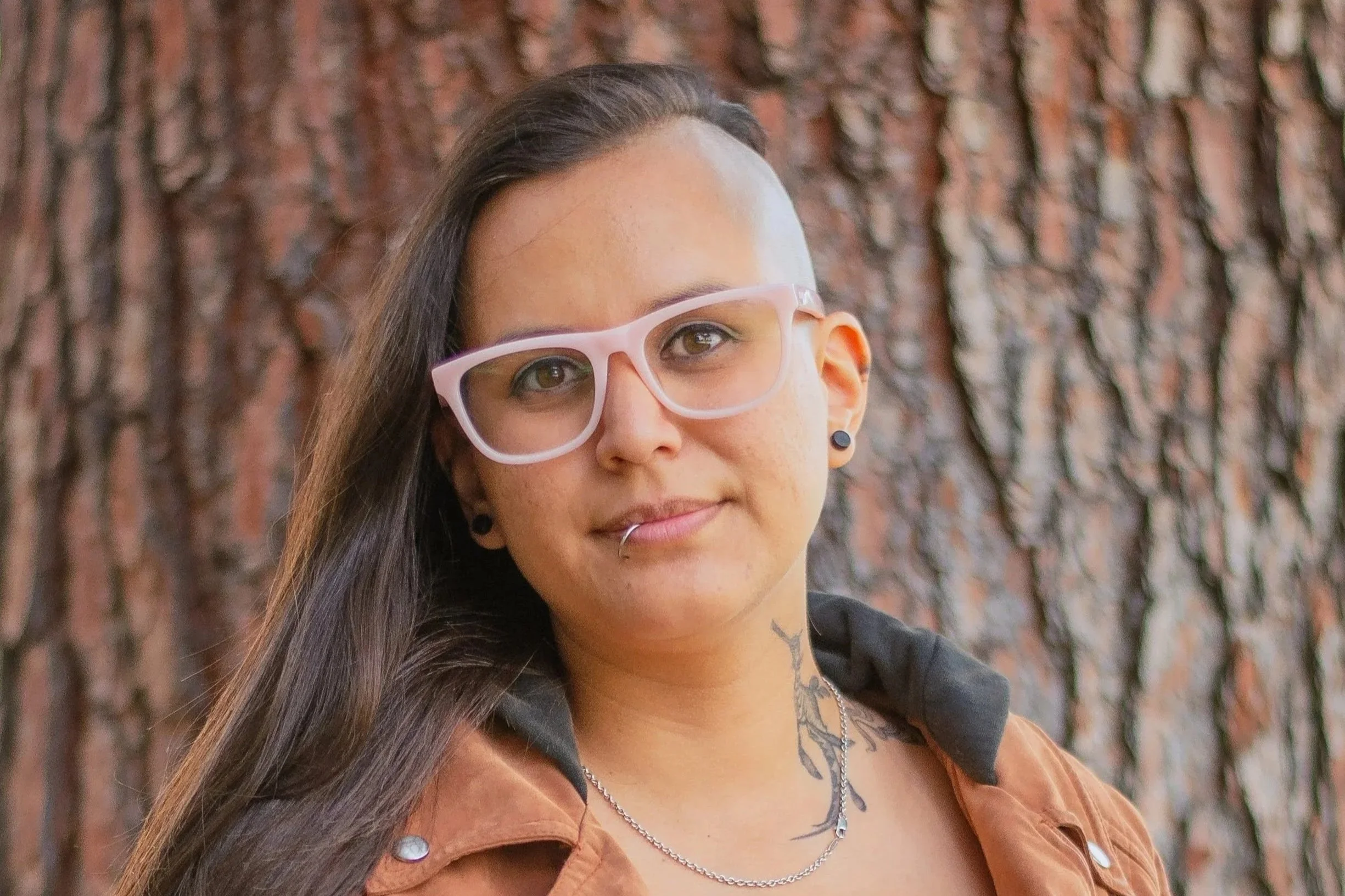 Close-up of therapist Cel Gonzalez, wearing pink glasses, earrings, and a necklace, standing in front of a tree trunk with textured bark.