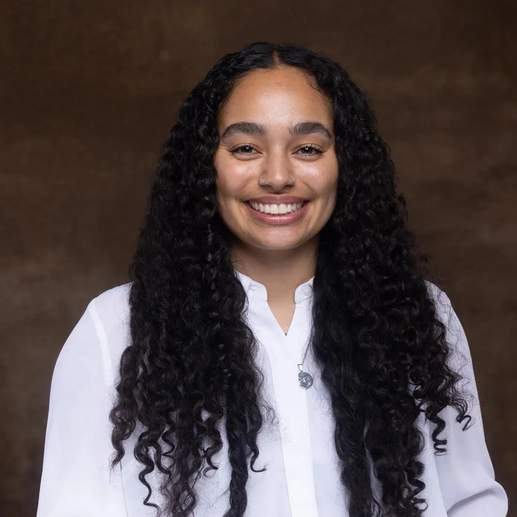 Therapist Elisa Rosado, has long, curly black hair, is smiling, wearing a white shirt, and standing against a brown background.