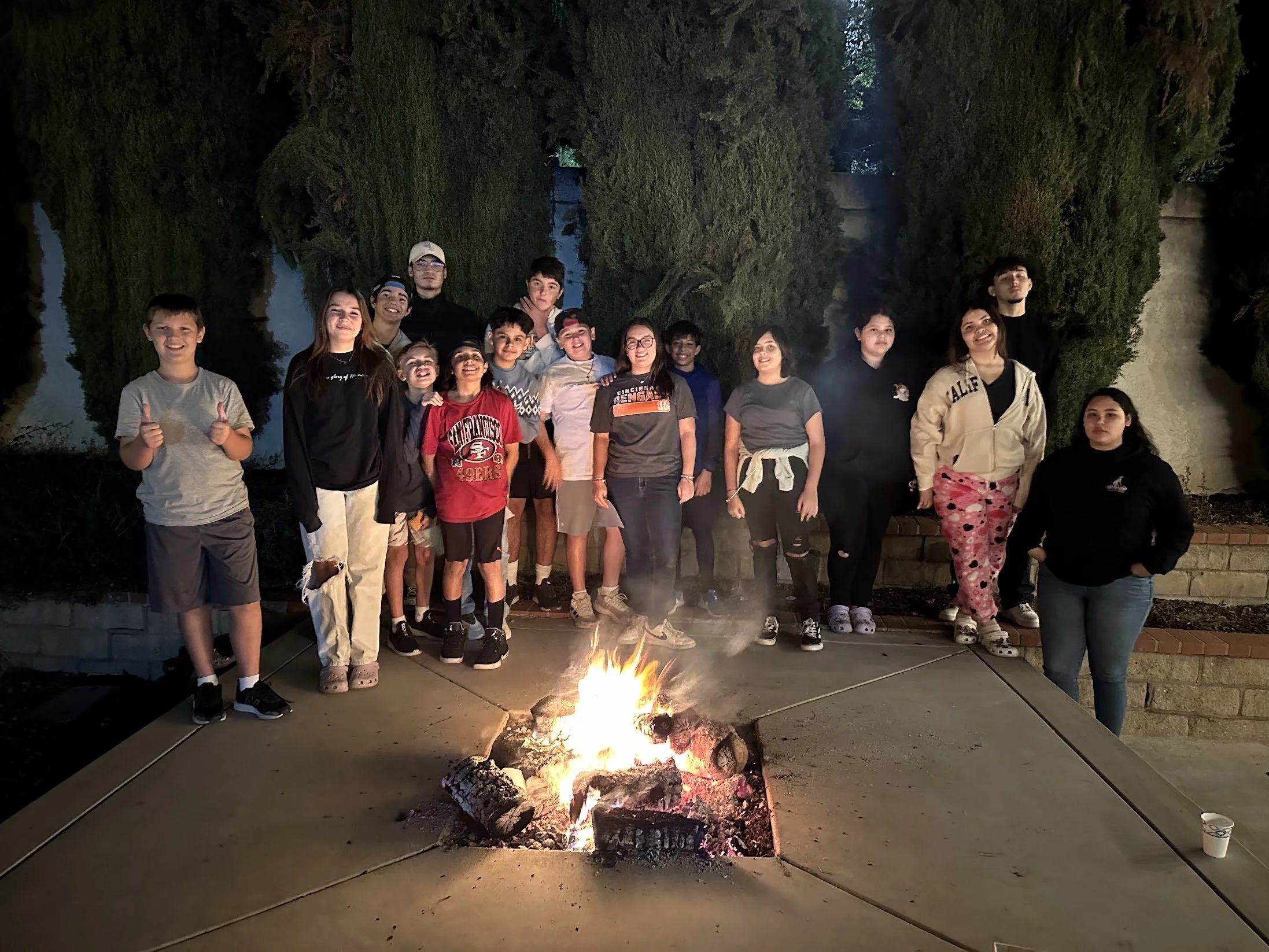A group of children and teenagers standing around a campfire at night outdoors, with trees and a water body visible in the background.