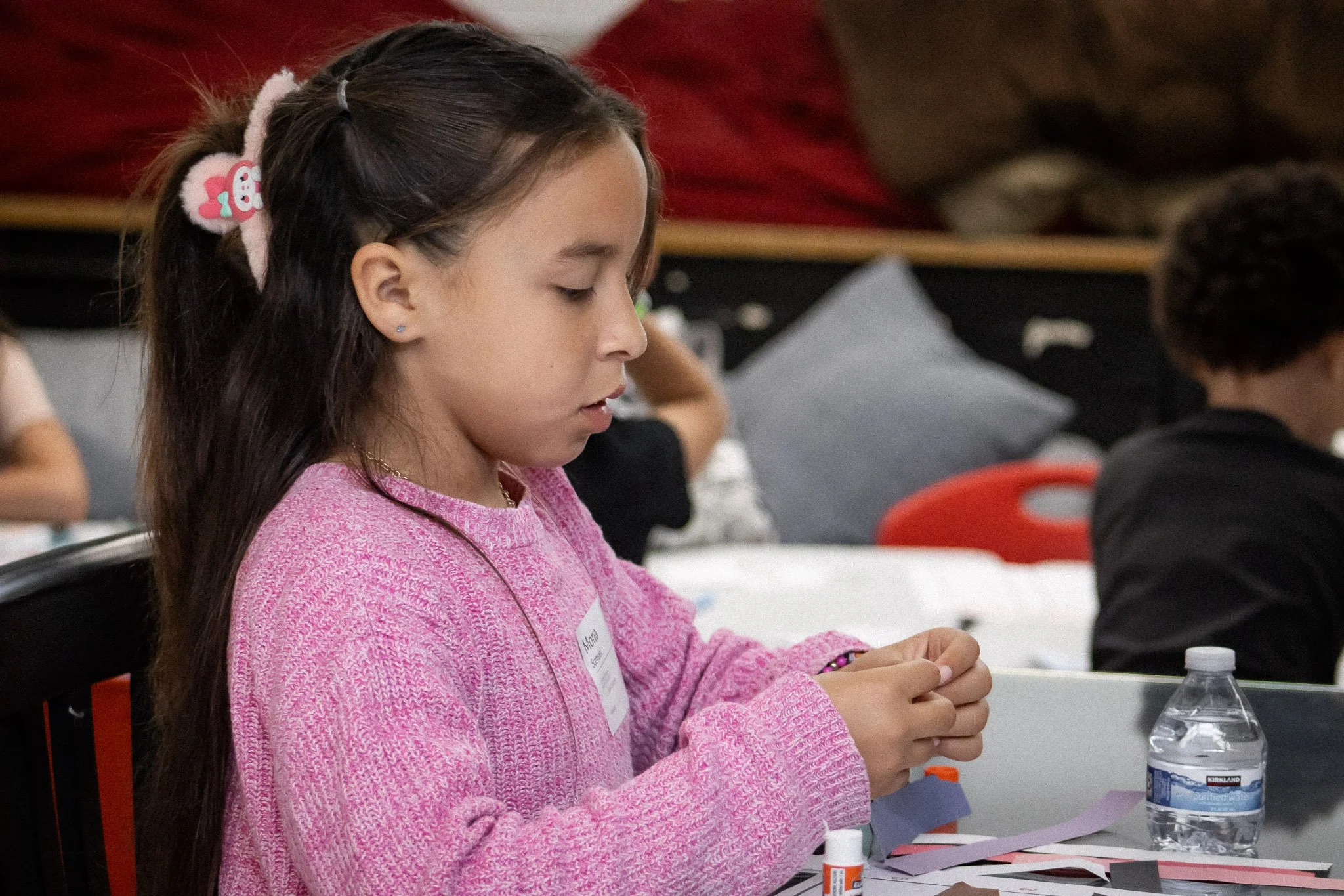 A young girl with long brown hair, wearing a pink sweater and a hair accessory, sitting at a table, working on an arts and crafts project with paper and glue, with a water bottle nearby.