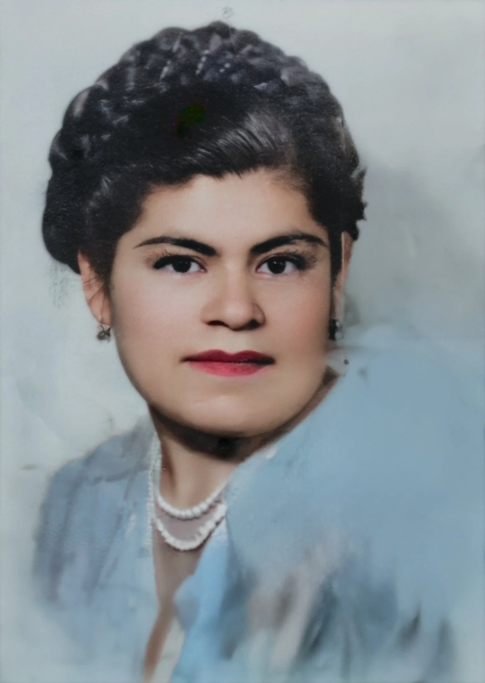 Portrait of a woman with dark hair styled in an updo, wearing earrings, a pearl necklace, and a light-colored blouse or dress.