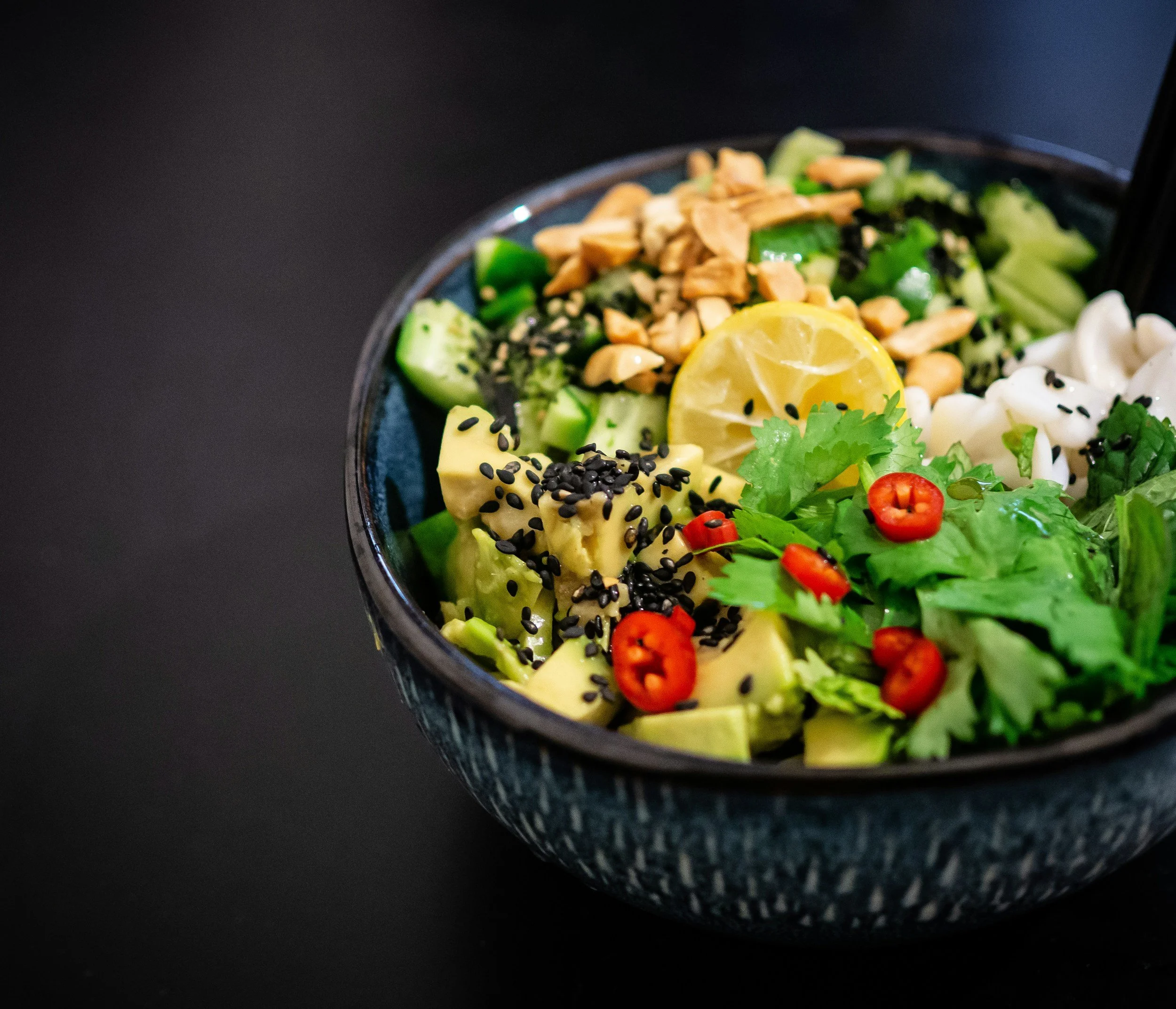 Close-up of an Asian-inspired salad bowl containing avocado slices sprinkled with black sesame seeds, chopped cucumbers, a slice of lemon, cherry tomatoes, chopped peanuts, and fresh herbs, on a black background.