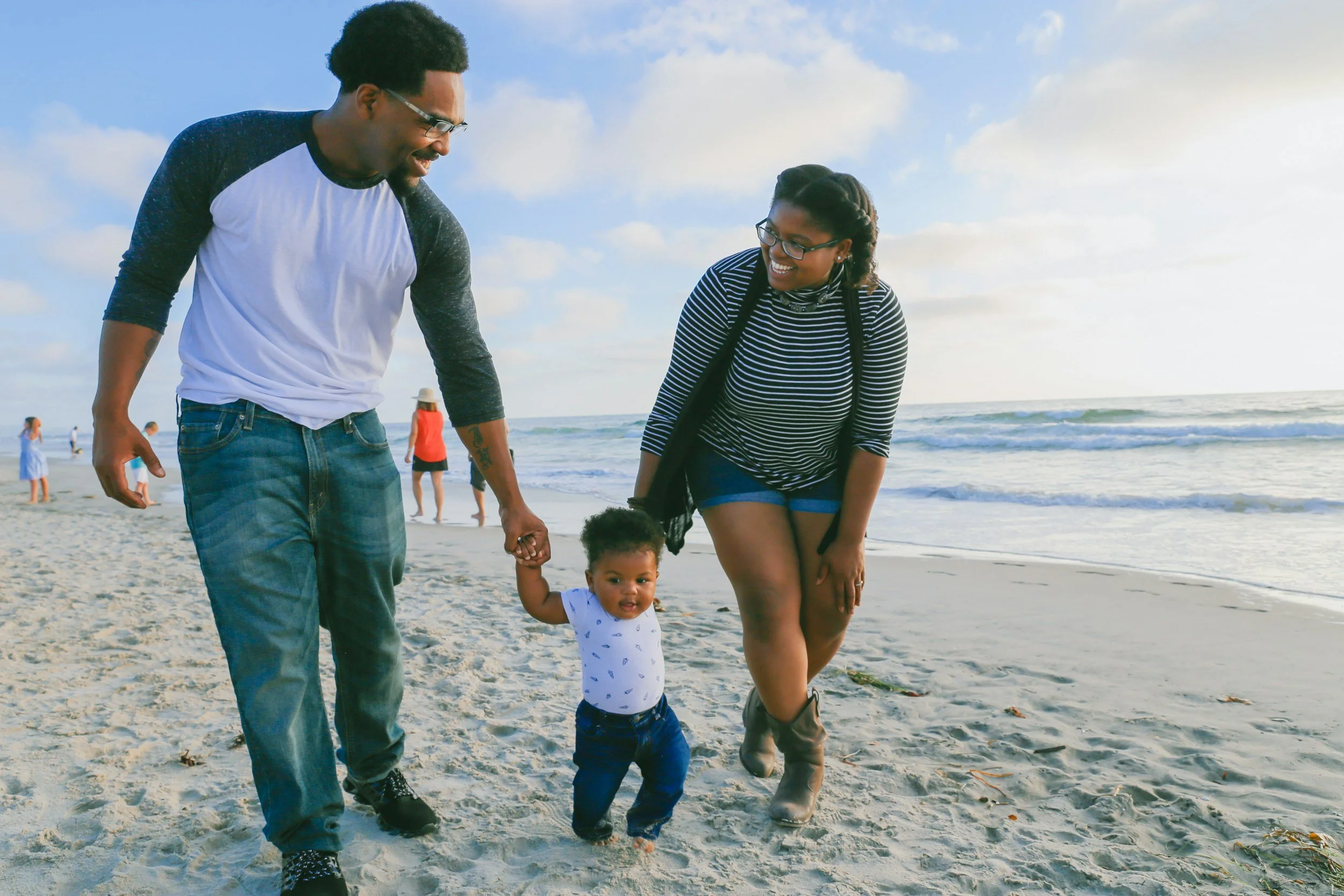 A family of three walking on the beach, holding hands. The father and mother are smiling, and their toddler is walking between them. The beach has other people in the background, and the sky is partly cloudy.