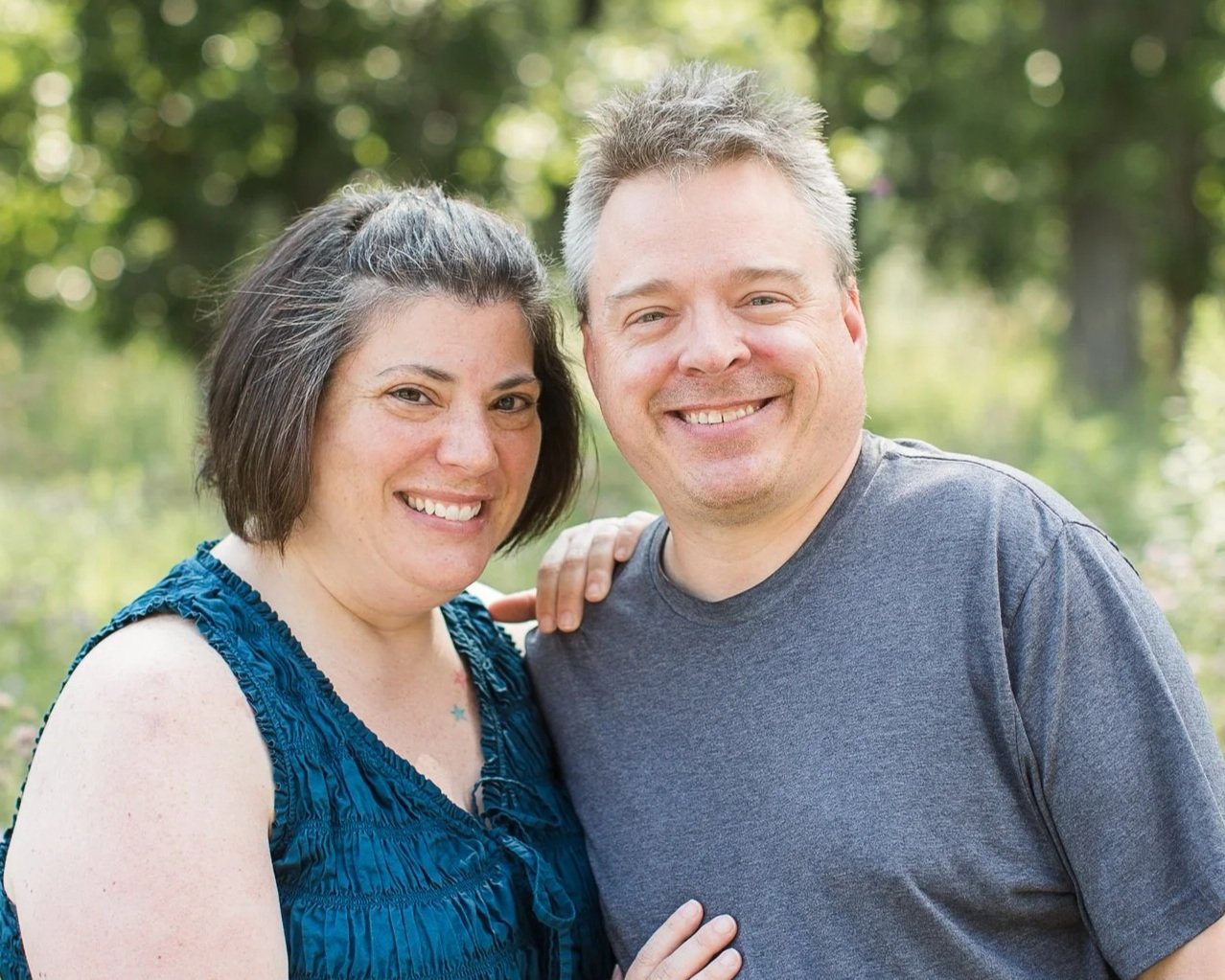 A smiling couple outdoors near a body of water, standing close together with the woman’s hand on the man's shoulder, surrounded by green trees.