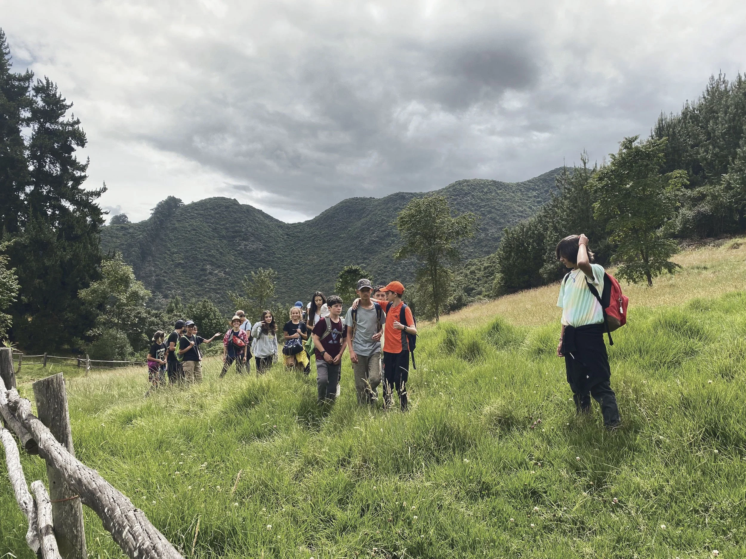 Grupo de personas caminando por un campo de hierba verde con montañas al fondo, cielo nublado, algunas personas llevan mochilas.