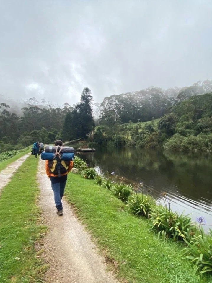 Persona con mochila caminando junto a un lago rodeado de vegetación, con cielo nublado.