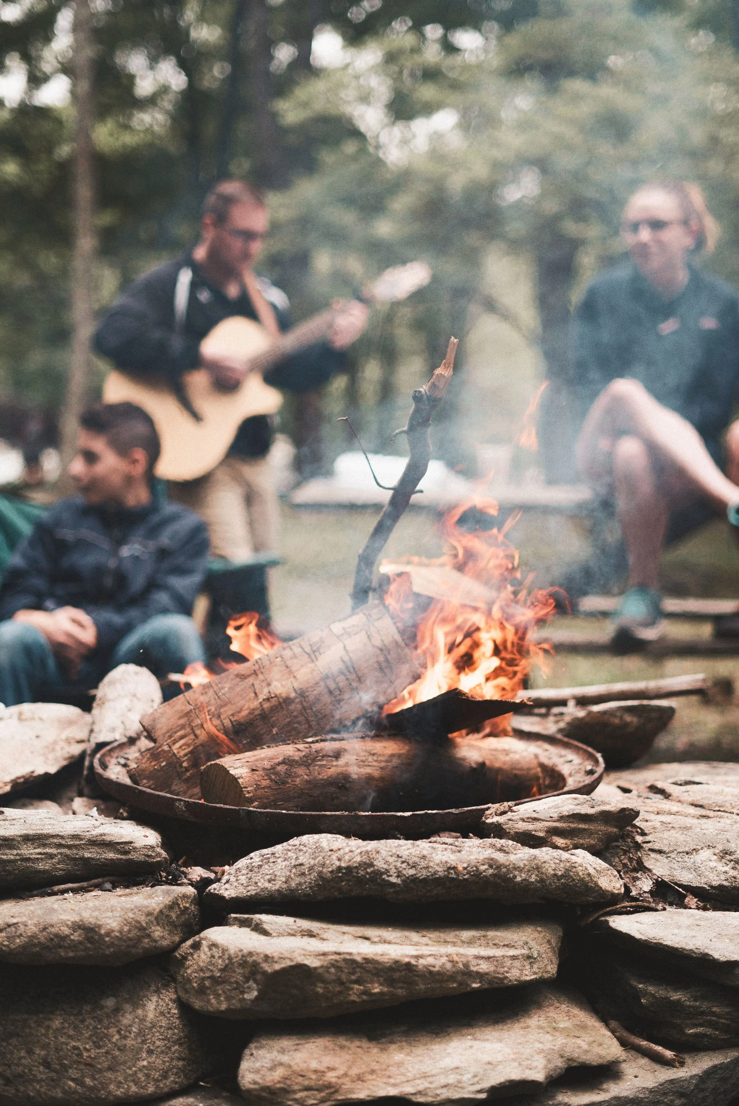 Personas alrededor de una fogata en el bosque, una tocando guitarra, otras conversando. Ambiente de campamento.