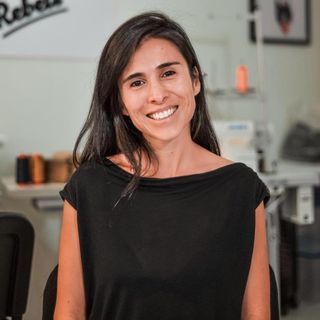 Persona sonriendo en un entorno de oficina, con equipo de costura al fondo.