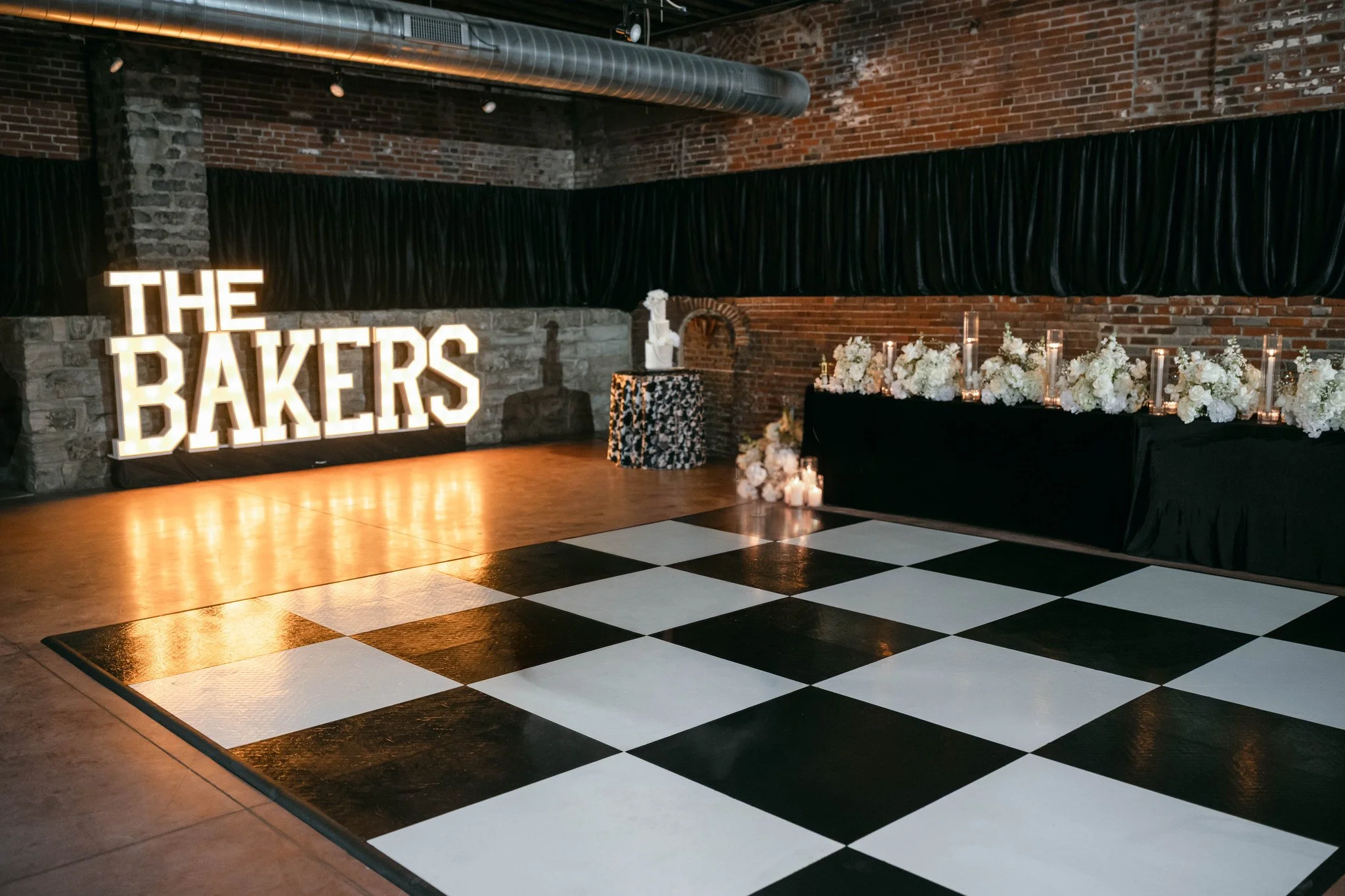 Indoor event space with black and white checkered flooring, brick walls, and a large illuminated sign reading 'THE BAKERS', decorated with white flowers and candles along a black table.