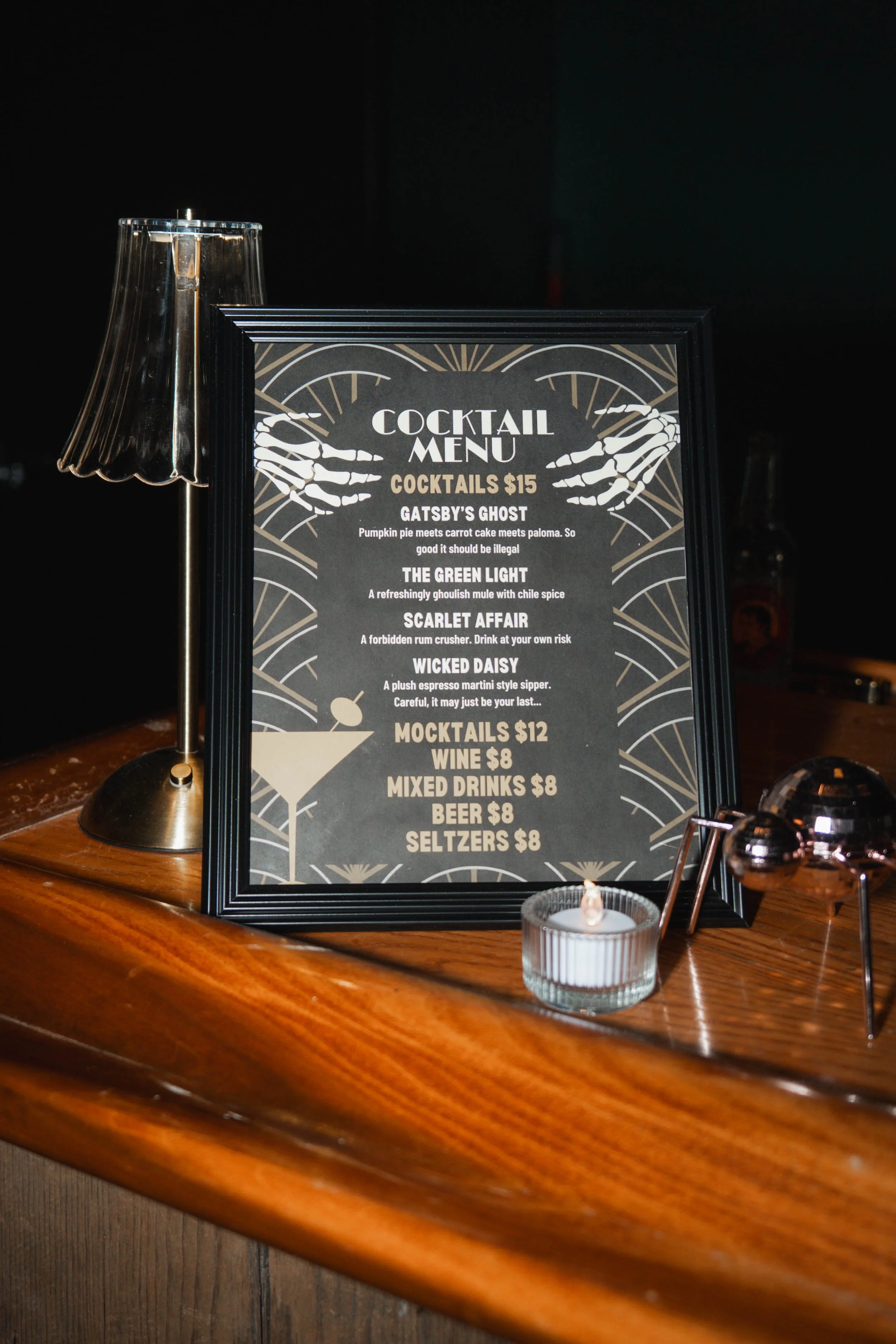 A framed cocktail menu on a wooden bar, with a small candle and metallic decor on the side.