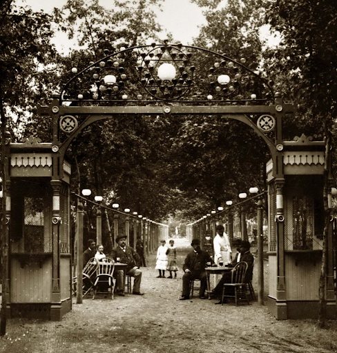 An outdoor scene at a park with a decorative archway for a tea garden entrance, with tables and chairs where people are seated and enjoying tea, surrounded by trees.