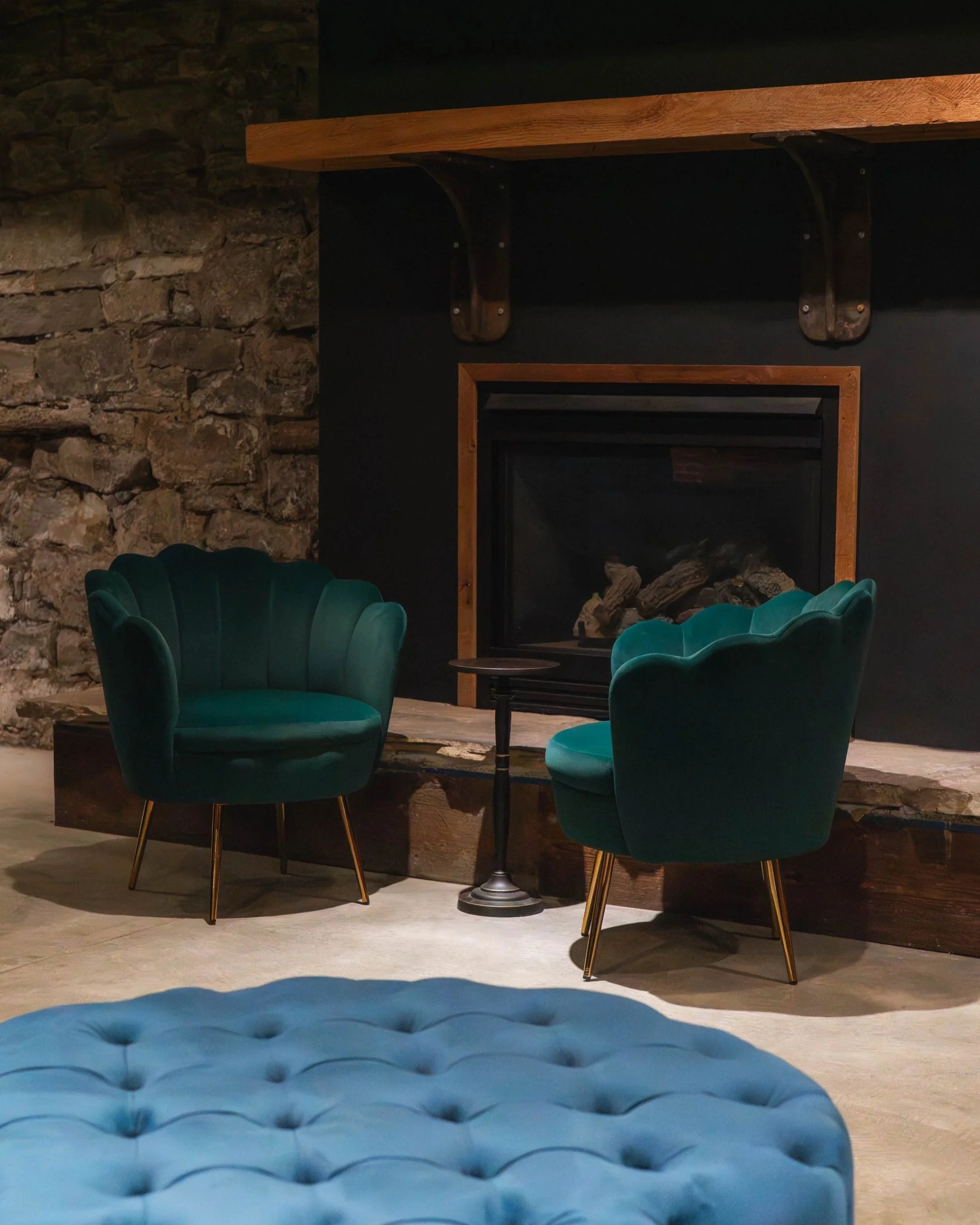 Two green velvet shell-shaped armchairs with gold legs are positioned in front of a fireplace with a stone wall on its left and a black wall with a wooden shelf above. A small black side table is between the chairs, and a blue tufted ottoman is in th
