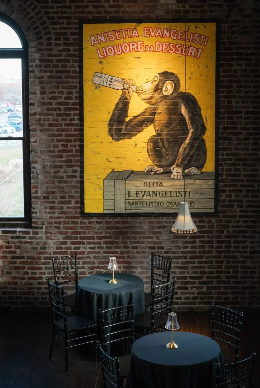Interior of a restaurant or cafe with exposed brick walls, a large colorful painting of a monkey drinking from a bottle hanging on the wall, and three black round tables with black tablecloths and small lamps. A window reveals parked cars outside.