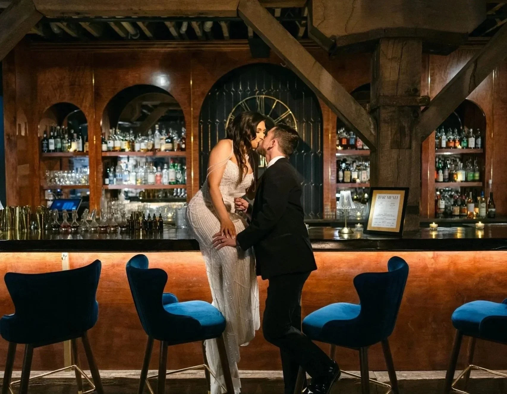 A couple kissing at a bar, with the woman sitting on the bar counter and the man kneeling in front of her. The bar has wooden shelves with bottles of alcohol in the background, and blue chairs are visible in the foreground.