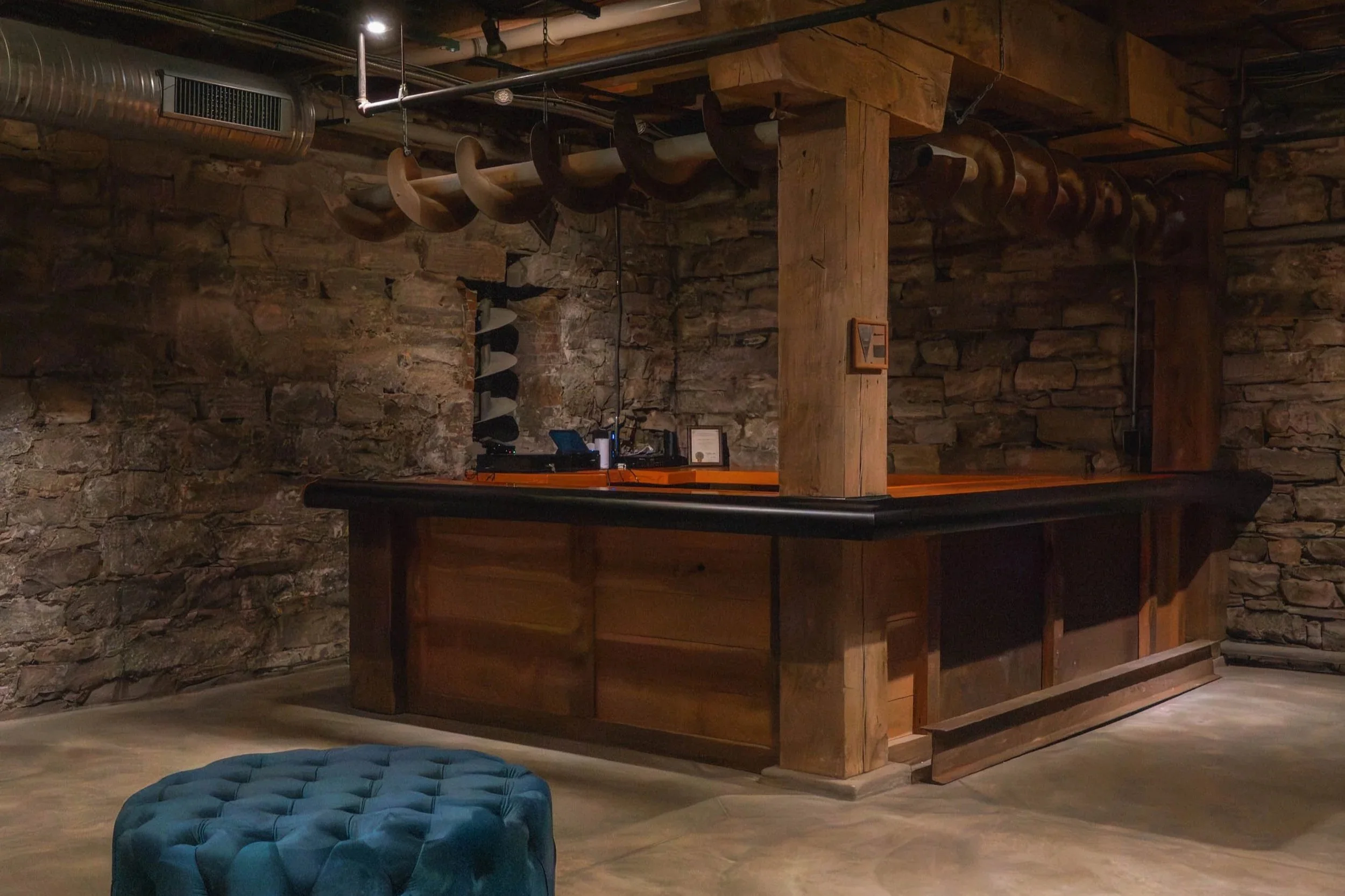 A rustic indoor space with brick and wood walls, wooden beams, and a bar counter. Decor includes a string of cow horns hanging from the ceiling. The foreground has a blue upholstered ottoman on a wooden floor.