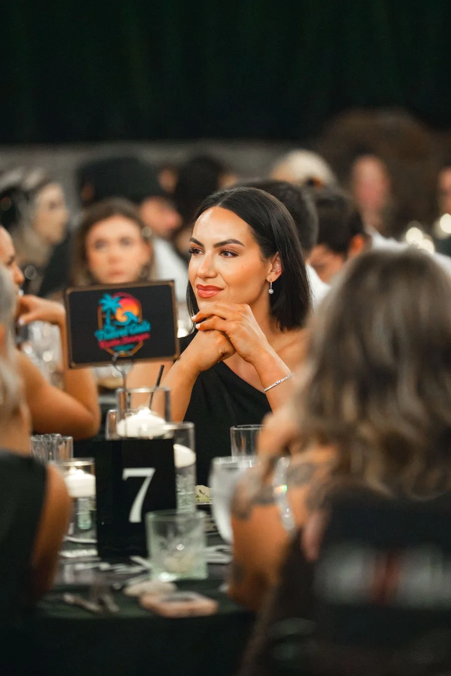 A woman with dark hair and earrings, sitting at a table during a social event, with her hands clasped under her chin, surrounded by other people.