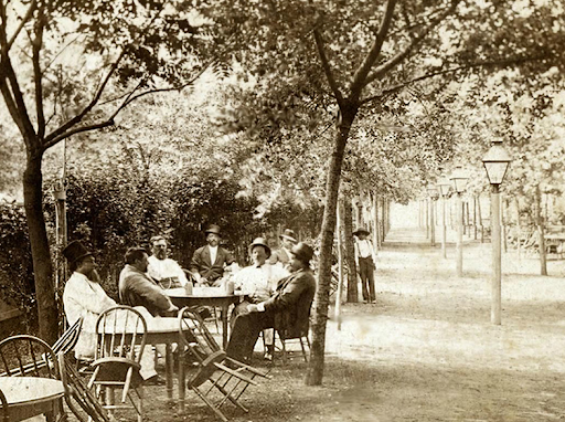 A group of people sitting at a table outdoors under trees, with a park pathway and lamp posts in the background.