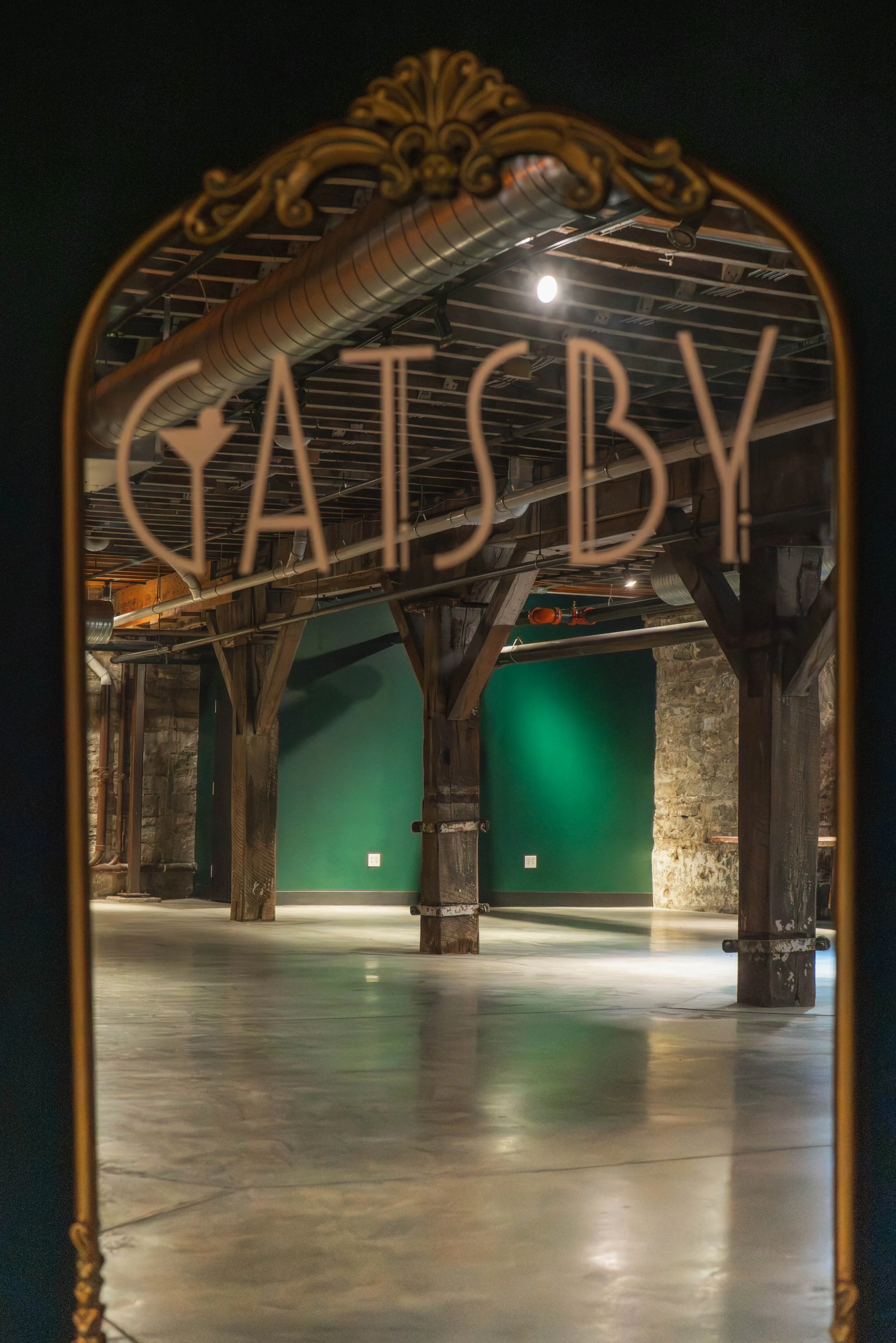 Empty industrial-style room with exposed wooden beams, brick walls, and green accent walls, viewed through a mirror with the word 'YARDSBY' on it.