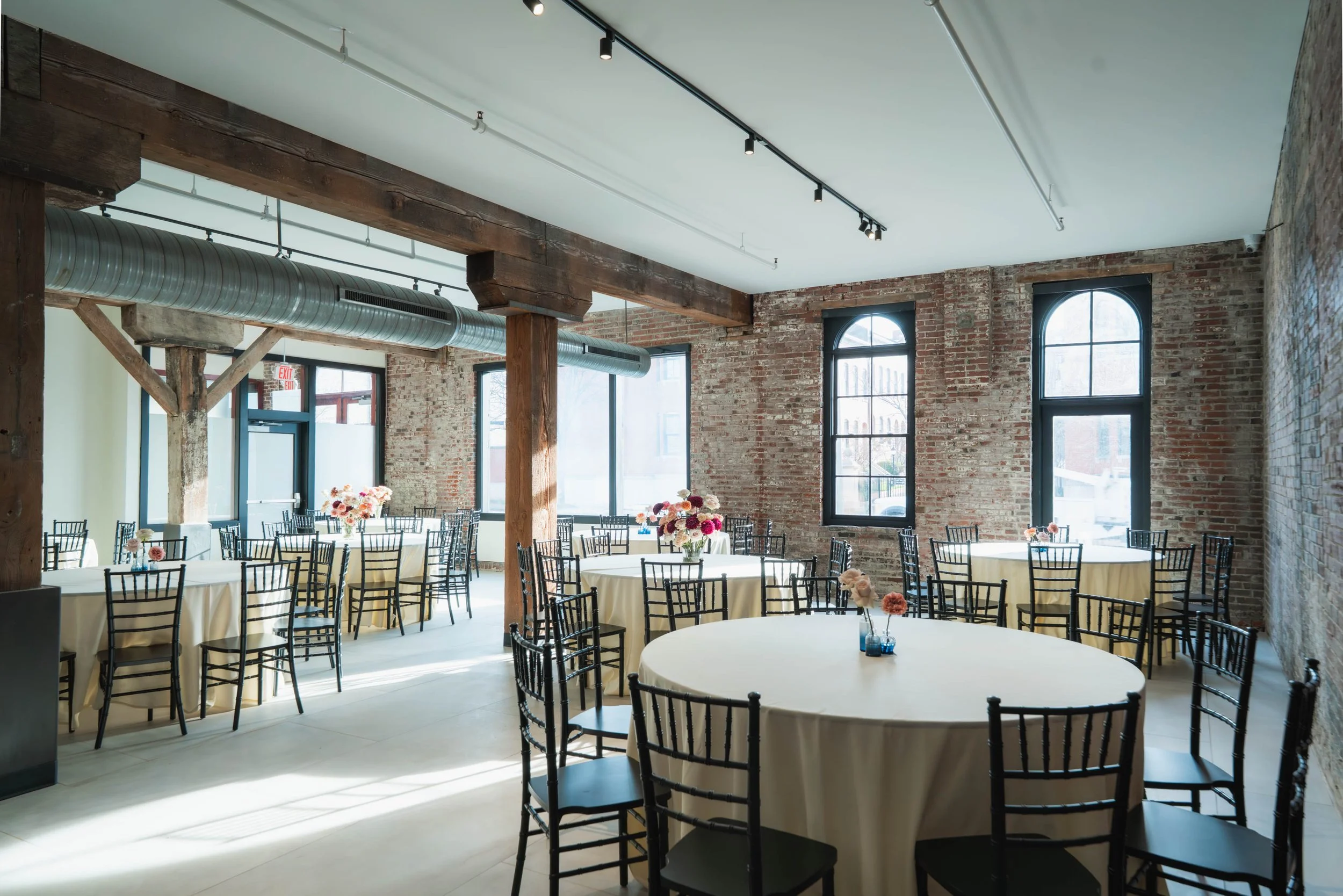 Event space with round tables covered in white tablecloths, decorated with small flower arrangements, and black chairs. Exposed brick walls, large windows, wooden beams, and industrial ductwork are visible.
